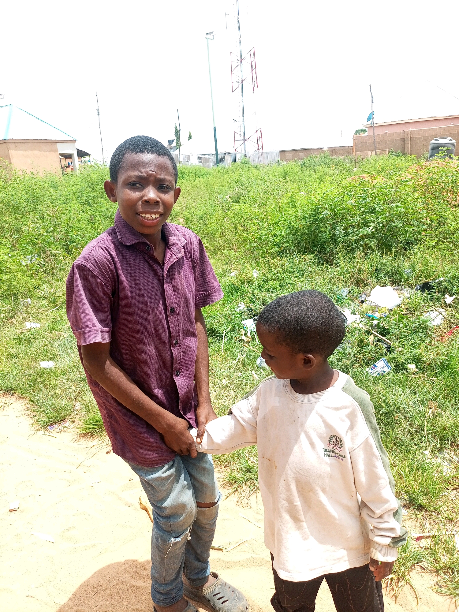 The image depicts two young boys outdoors in what appears to be a semi-urban or rural environment in Tempure, Nigeria. The boy on the left, with dark skin and short hair, is wearing a short-sleeved, collared, dark red button-up shirt with a subtle checkered pattern and light blue distressed jeans. He is looking directly at the camera with a wide smile, revealing his teeth. The boy on the right, also with dark skin and short hair, is wearing a light-colored long-sleeved sweatshirt with green accents on the sleeves and collar, and dark pants. He is turned slightly to his left, looking down and to the side, and is holding the hand of the boy on the left.
The environment is characterized by dry, sandy ground in the foreground, transitioning to overgrown green grass and vegetation in the background. Scattered among the vegetation are various pieces of white and dark-colored trash, including plastic bags and wrappers. In the background, there are several buildings, including a light-colored structure with a blue roof on the left, and a brick building with a water tank on the right. A tall communication tower with red and white lattice structures is visible in the far background, suggesting proximity to infrastructure. The sky is bright and overcast. The overall scene appears to be a candid moment captured outdoors, possibly in a less maintained area.