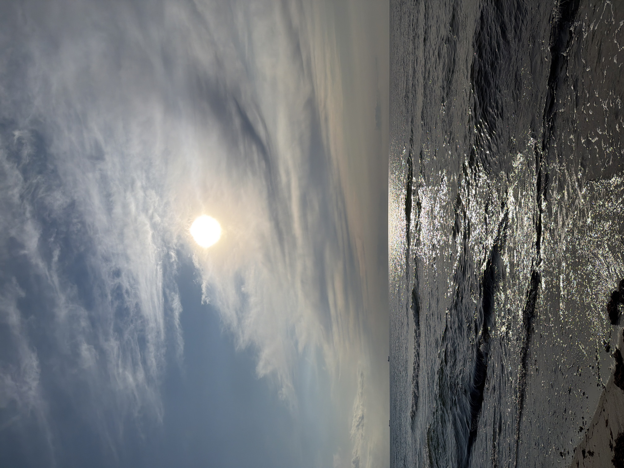 The image depicts a serene seascape under a cloudy sky. The sun, a bright orb, is partially obscured by wispy clouds, casting a shimmering reflection on the water's surface. Gentle waves lap at the sandy shore, with foam and seaweed scattered near the water's edge. In the distance, two small ships are visible on the horizon. The overall impression is one of tranquility and natural beauty. The scene is likely located in Baltiysk, Russia, a port city on the Baltic Sea.