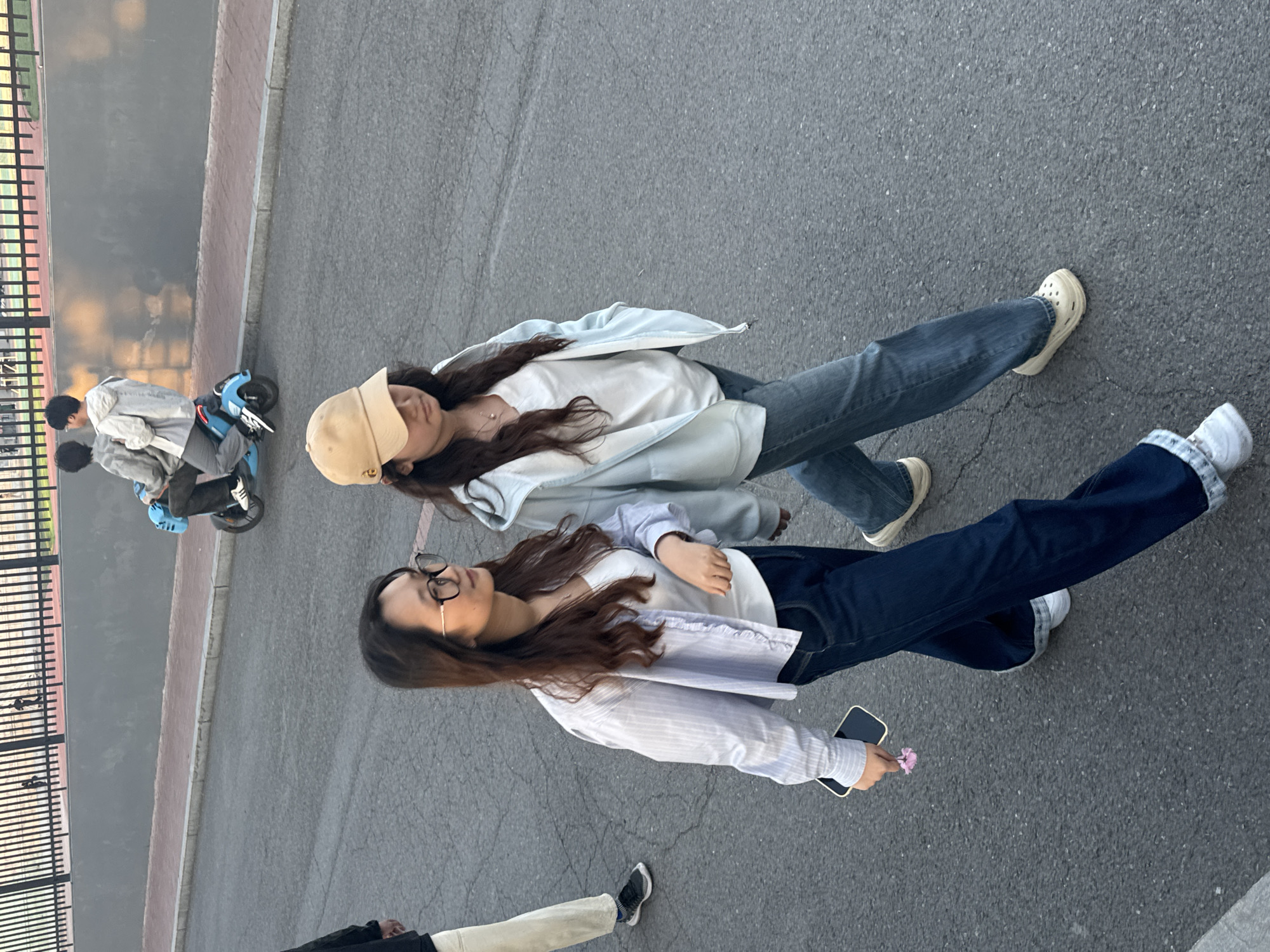 Two young women walk along a gray asphalt surface. One woman wears glasses and a long-sleeved striped shirt, dark jeans with white socks, and holds a cell phone and a small flower. The other woman wears a tan baseball cap, a white shirt, a light blue jacket, and blue jeans. Nearby, two individuals sit on a blue electric scooter. A black fence runs along the edge of the paved area. The location is presumed to be in Zhongtun, China.