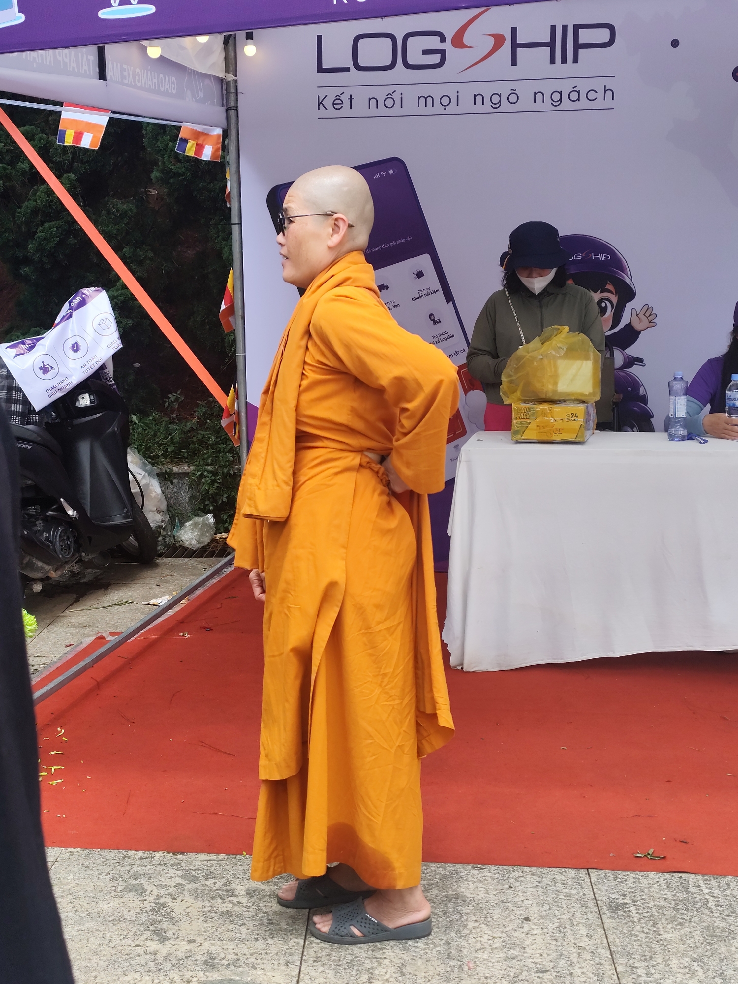 The image, likely taken in Da Lat, Vietnam, features a bald individual dressed in an orange monastic robe and dark glasses, standing on a red carpet that appears to be part of an event setup. The individual has one hand on their hip. Behind them, there's a white table with two individuals present. One is wearing a dark hat and a facemask. On the table, there's a yellow package and two water bottles. In the background, the "LOGSHIP" logo is visible, indicating that the event might be promotional for this service. There is a black scooter and a white bag to the left, and a tree behind the scooter. Flags hang above.