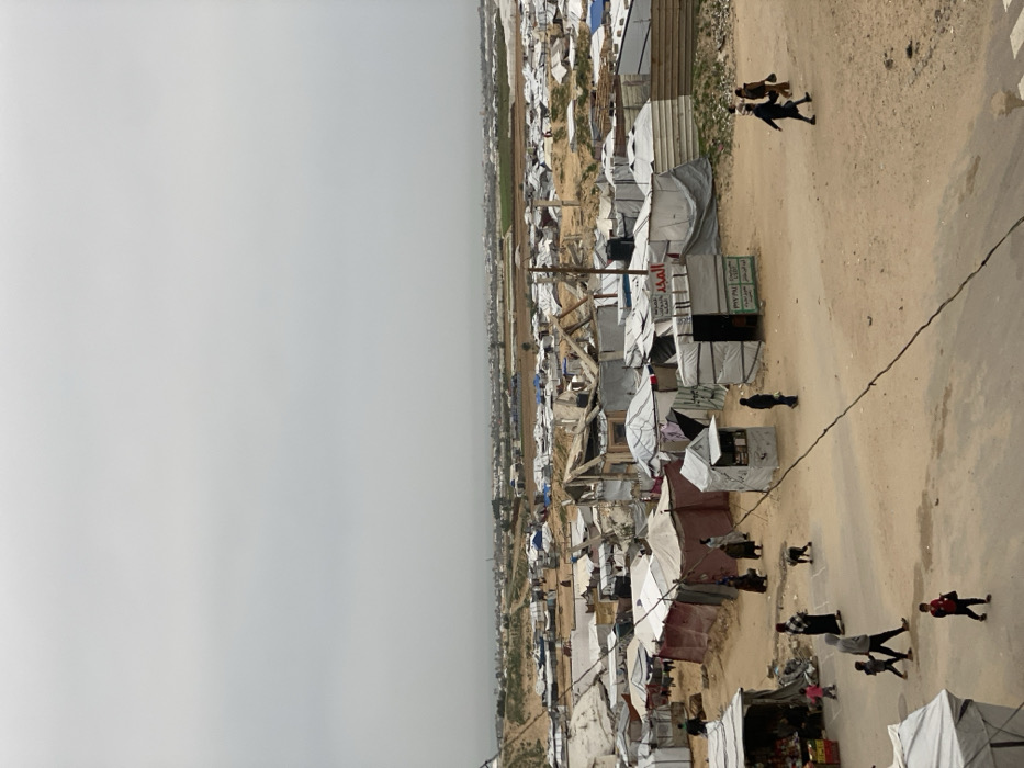 An overhead view depicts an expansive informal settlement or refugee camp located in Khan Yunis, Palestinian Territory, under an overcast sky. The scene is dominated by numerous densely packed, makeshift shelters constructed primarily from white, grey, and some reddish-brown tarpaulins stretched over visible wooden and metal frames. Several structures exhibit signs of damage, with exposed supports.
The ground consists of light-brown, dusty earth, forming irregular paths between the shelters. In the mid-ground and background, the settlement extends broadly, eventually transitioning into distant patches of green agricultural fields and sparse buildings on the horizon.
Approximately two dozen individuals, including adults and children, are visible navigating the paths and open areas. They are walking individually or in small groups. In the mid-ground, a child is observed kneeling near a small, dark animal.
Specific structures include small commercial kiosks: one on the right features Arabic text "المجد" (Al-Majd) and "PAY PAL" signage, while another on the left functions as a market stall displaying produce in crates. Black water tanks are present on the roofs of some shelters. A dark cable or line diagonally crosses the lower-center portion of the image.