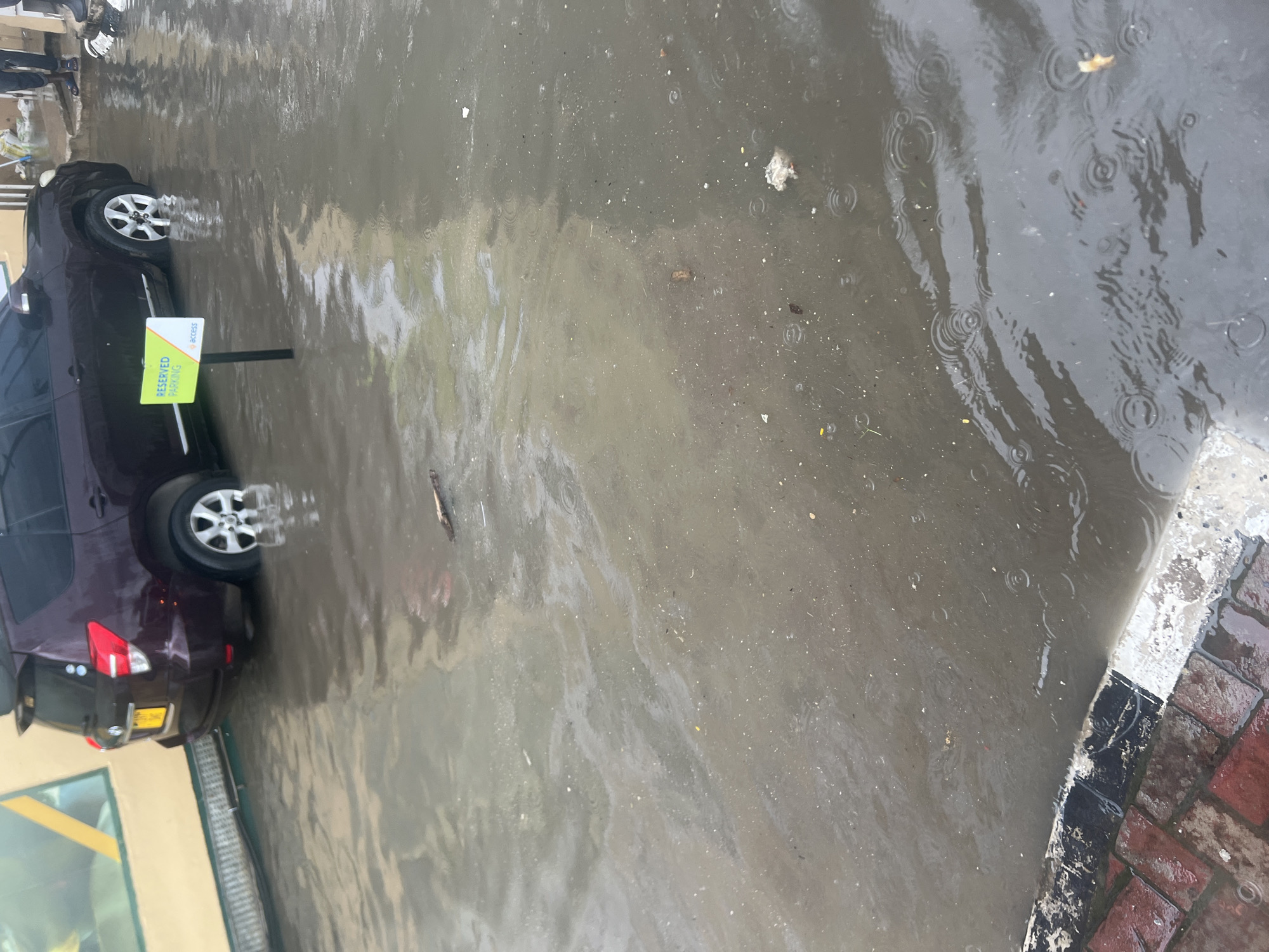 The image captures a street or parking area in Dar es-Salaam, Tanzania, significantly flooded with murky rainwater. A dark-colored, possibly burgundy, car (appearing to be an SUV or hatchback) is parked on the left side, with its wheels and lower body partially submerged in the standing water. Next to the car, a yellow-green sign on a black pole reads "RESERVED PARKING" in white text, with a white "ACCESS" logo above it.
The surface of the floodwater is disturbed by numerous concentric ripples, indicating that rain is actively falling or has very recently fallen. Various small pieces of debris, including what appears to be litter and natural matter like twigs or leaves, are visible floating on the water. On the right side of the frame, a red brick sidewalk or curb is visible, wet and stained, with a painted black and white stripe along its edge, separating it from the flooded street. The scene suggests an urban environment experiencing the effects of heavy rainfall and subsequent flooding during the daytime, likely under an overcast sky.