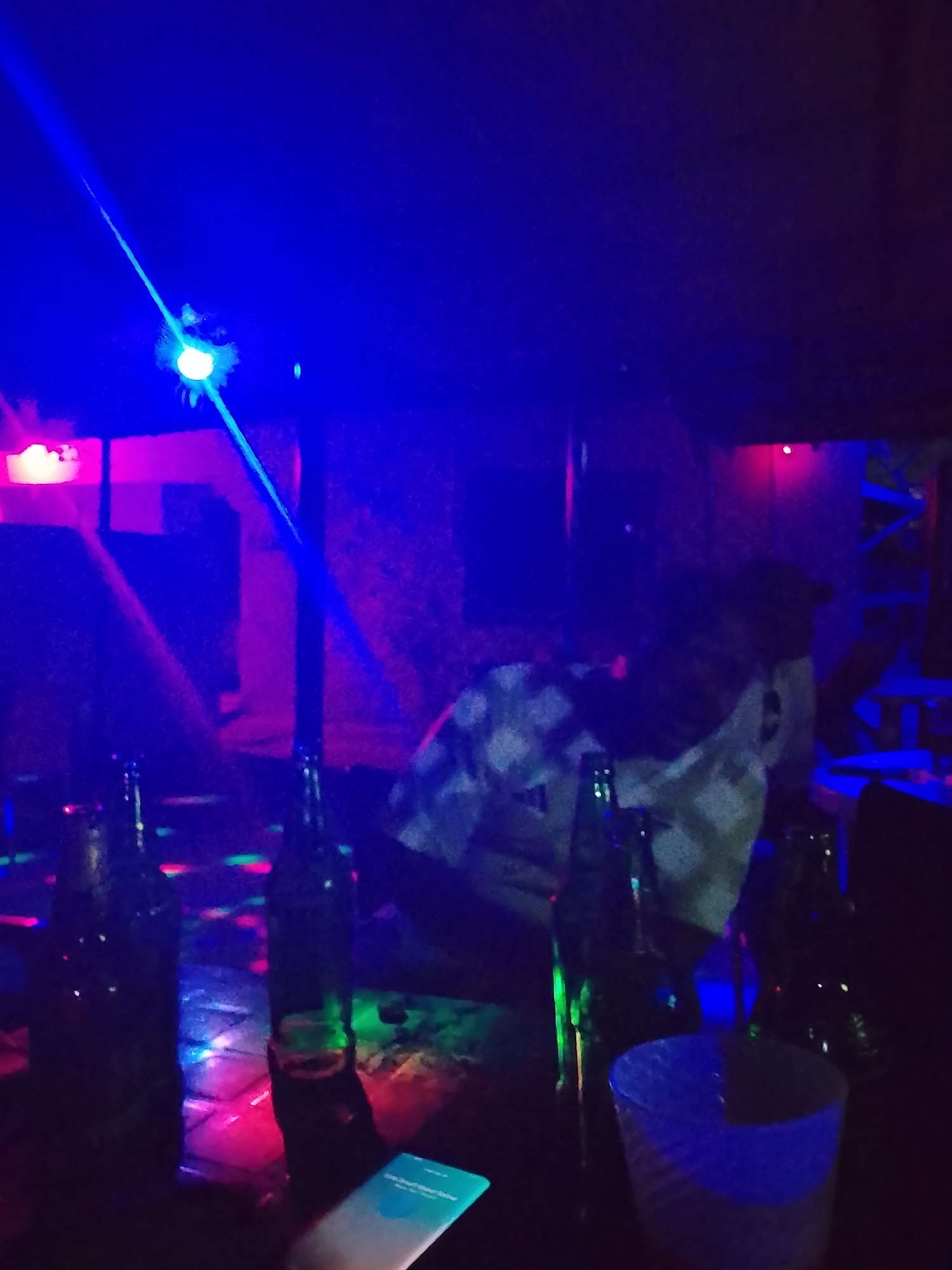 This media file depicts an indoor, low-light setting, likely a bar, club, or party, illuminated by vibrant artificial lights at night. A dominant blue light beam diagonally crosses the upper left portion of the image, emanating from a bright light source in the corner, possibly a disco ball or projector.
In the mid-right, a person is partially visible, appearing to be seated or leaning. They are wearing a light-colored shirt with a subtle pattern, their features obscured by the dimness and colored lighting.
Numerous dark glass bottles, resembling beer bottles, are scattered across a surface in the foreground and mid-ground. This surface, possibly a table, reflects various colored light patterns—including green, red, and blue dots—characteristic of disco or party lighting. A light-colored cup, appearing to glow blue from the ambient light, is positioned in the lower right. In the lower-middle foreground, a smartphone with a brightly lit screen is visible, displaying what appears to be a notification or a search bar, with "Search your phone" faintly legible.
The scene suggests a social event or recreational activity, typical of nightlife environments. The strong, colored lighting and visible elements like bottles and a laser beam contribute to a festive and energetic atmosphere. The time of day is clearly night due to the reliance on artificial light.