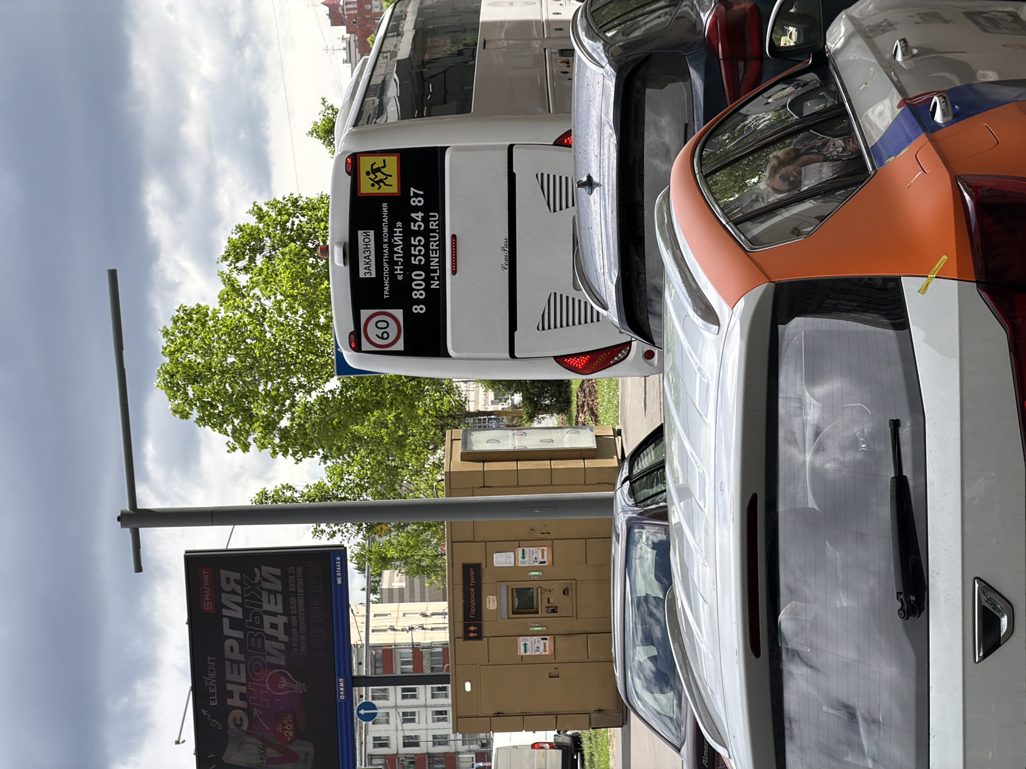 The image captures an urban street scene, likely in Unknown, Unknown, with a mix of vehicles and urban infrastructure. A bus is prominently featured, with clear labeling including a speed limit of 60 km/h and contact information for "N-LINE", and its rear. Beside the bus, several cars are parked in the lane, featuring a vehicle with an orange and white color. An advertisement pillar stands nearby, marked with Russian text, indicating "ENERGY OF NEW IDEAS". A municipal toilet facility is placed next to cars, suggesting a public space. The sky, overcast, contrasts with the greenery of trees in the background.