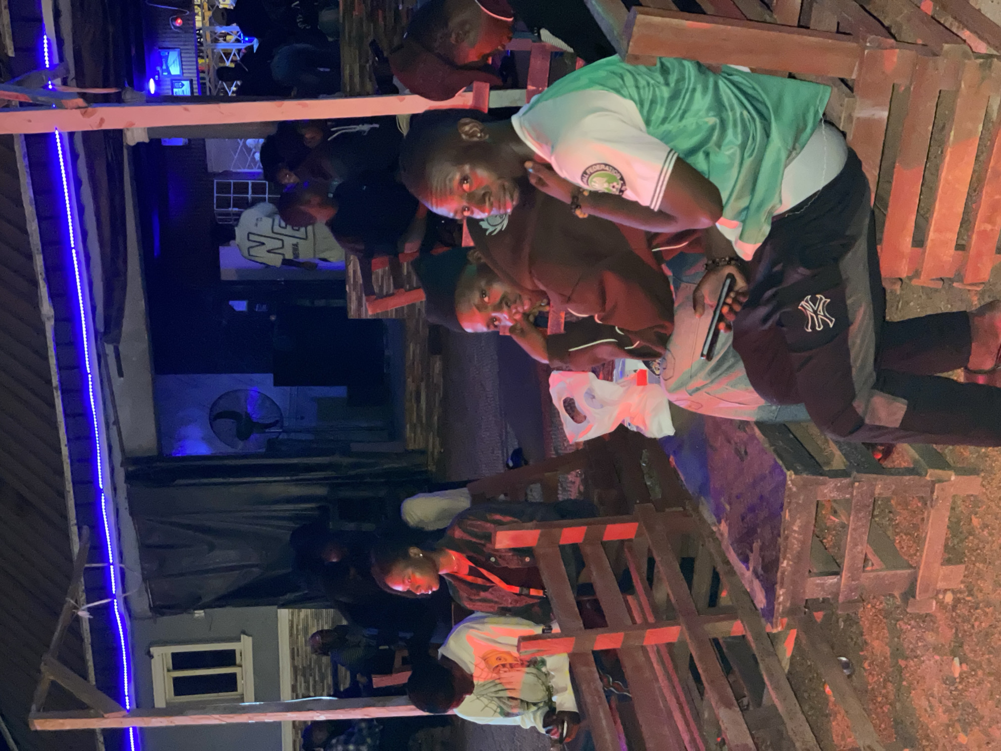 The image depicts a group of young Black men gathered outdoors at night, likely in Durumi, Nigeria. The setting appears to be a casual, informal gathering space with wooden seating structures, possibly a makeshift bar or lounge area. Red and blue lights illuminate the scene, suggesting a lively atmosphere, possibly a party or social event.
Several individuals are visible, and some are looking towards the camera. One person in the foreground is wearing a white t-shirt with a graphic print and a dark jacket, while another is sporting a green jersey with white stripes. Another man is wearing a dark brown top. The men are interacting with each other and seem relaxed and engaged. One person is holding a smartphone.
The ground is covered with gravel or dirt. In the background, there are glimpses of buildings and structures, including a window and what looks like an air conditioning unit or fan. The overall impression is one of a communal gathering in a somewhat rustic, outdoor environment at night. The lighting creates a vibrant and energetic mood.