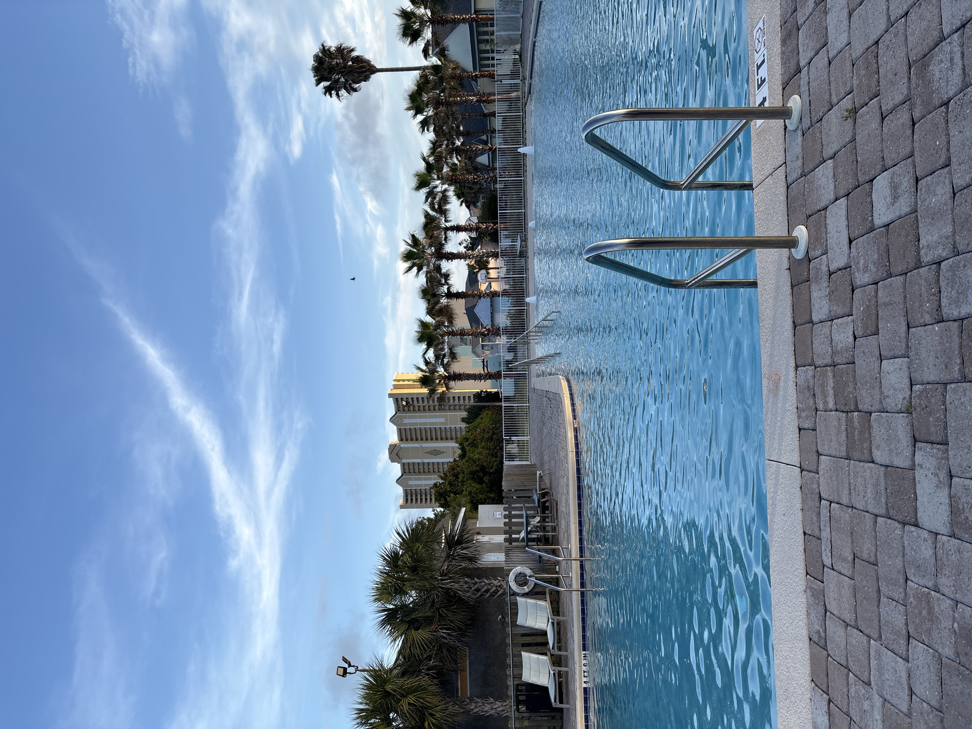 The image depicts a swimming pool area with a view of palm trees and buildings, likely in Panama City Beach, Florida. The pool water is light blue, and there are metal railings for entry and exit. The pool deck is made of brick pavers. Palm trees line the edge of the property, and tall buildings are visible in the background. The sky is light blue with scattered clouds.