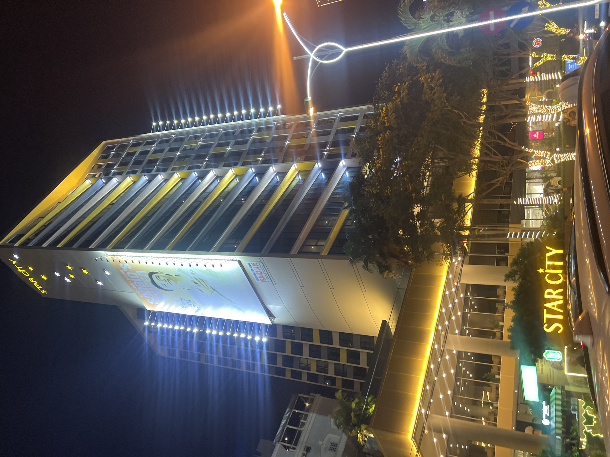 The image shows a building at night, likely located in Nha Trang Ward, Vietnam. The building, named "Star City," is tall and modern, with a facade featuring a large advertisement and distinctive yellow and silver stripes. Numerous lights illuminate the building's exterior, creating a bright glow. Streetlights and decorative lights further illuminate the surrounding environment. A vehicle is visible in the foreground, possibly indicating a street scene. The presence of trees and street signs suggests an urban setting.