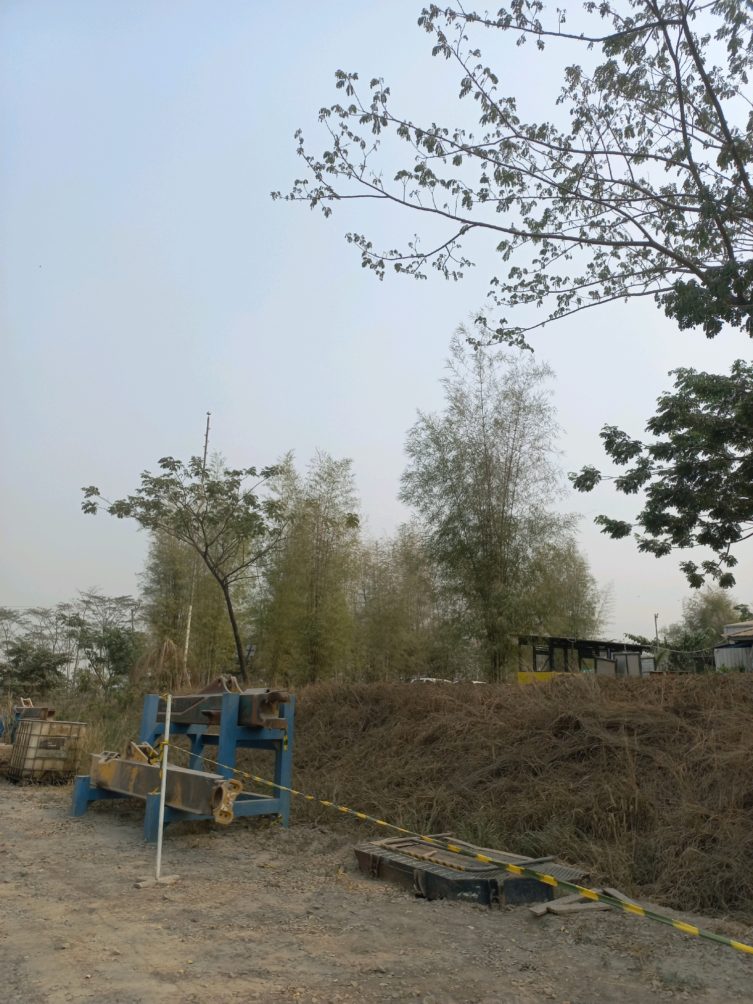 This is a daytime, eye-level, outdoor, wide-angle photograph of a construction or storage site, likely located near Paringin, Indonesia. The sky is overcast and pale. In the foreground, there is a dirt path with loose gravel. To the left, a blue and yellow metal structure, possibly part of construction equipment, is positioned on the ground. A white pole with a yellow tape is also present. Further back, a large pile of dry, brown vegetation or debris forms a mound. A yellow and black striped caution tape runs across the scene, partially obscuring a black, metallic object on the ground. In the midground, there are several tall, thin bamboo trees and a larger tree with sparse green leaves. In the background, there are some simple structures with dark roofs, possibly sheds or shelters, and more vegetation. The overall impression is of an undeveloped or work-in-progress area.