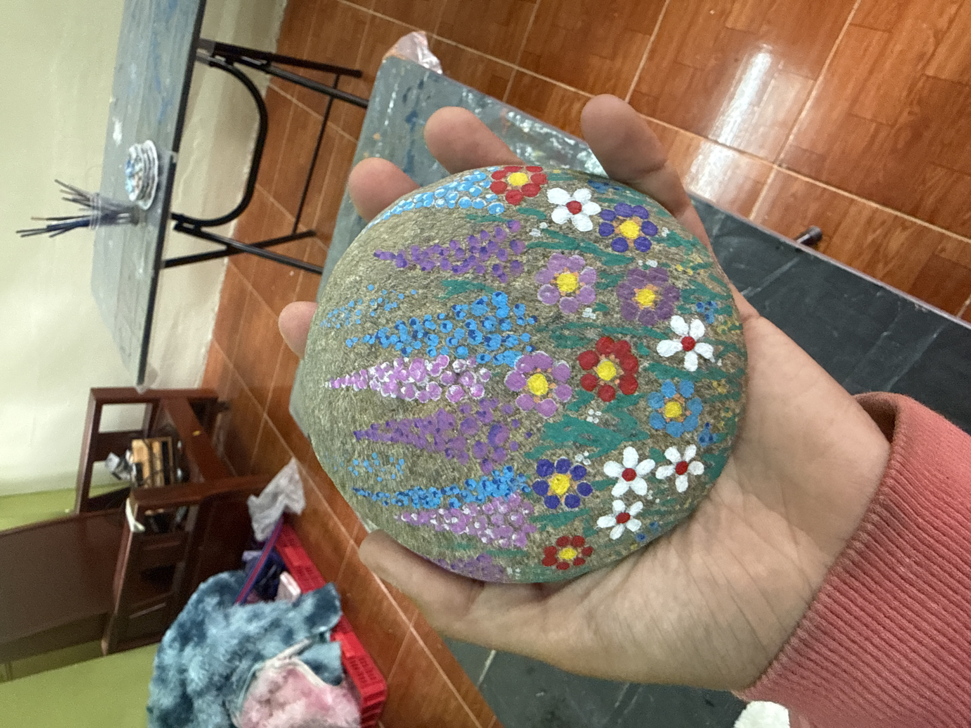In Đạ Cháy, Vietnam, a person's hand, covered by a pink sweater sleeve, holds a round, grey stone painted with a colorful floral design. The stone features rows of lavender and blue flowers interspersed with smaller red, white, and purple flowers with yellow centers. Green stems and leaves fill the background of the painting. In the background, a wooden chair with a stack of items is visible, along with a red plastic crate containing blue and pink fluffy blankets. A folding table with a dark surface holds a clear cup filled with paintbrushes and a stack of small, circular objects. The floor is tiled with a reddish-brown pattern.