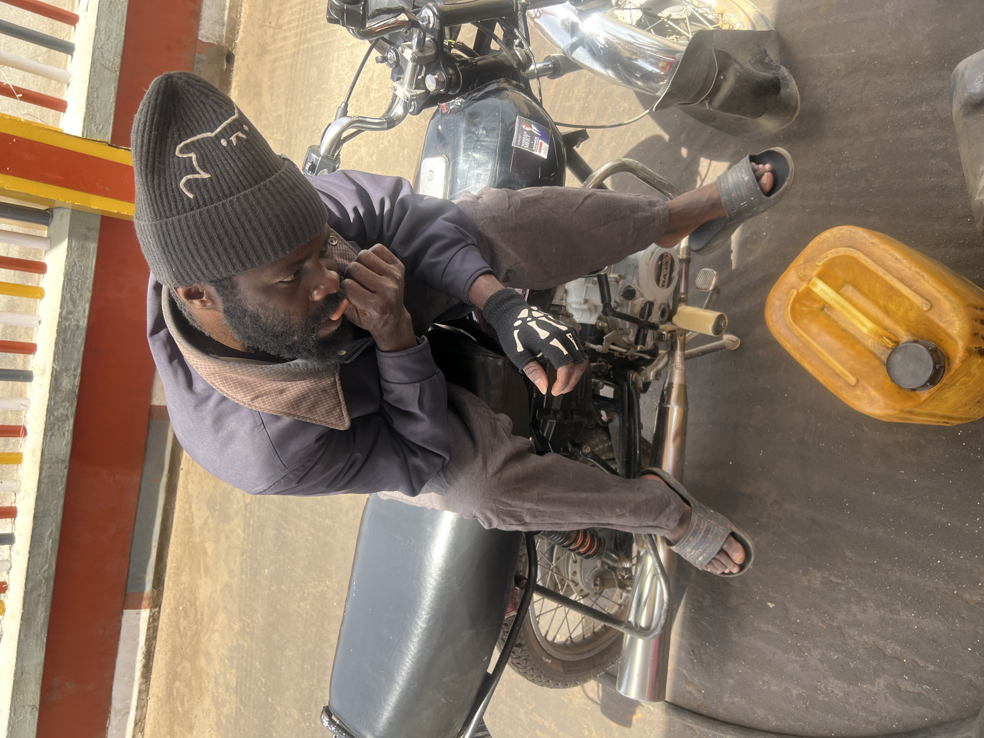 An overhead shot shows a man seated on a motorcycle in Jos, Nigeria. He wears a black beanie with a white graphic, a grey jacket, brown pants, black gloves with a white bone design, and grey sandals. The motorcycle is black with chrome accents. A yellow fuel container sits next to the motorcycle. The scene is set on a concrete surface next to a painted barrier.