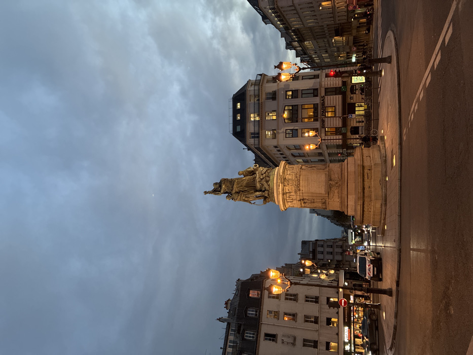 A Parisian roundabout features a statue atop a stone pedestal, dominating the scene at dusk. Classic architecture flanks the street, illuminated by vintage-style lampposts. A BNP Paribas bank branch is visible on one building. The cloudy sky casts a subdued light. A crescent moon is barely visible. Street markings and a white police vehicle mark the street. Traffic lights glow red, adding to the city's ambiance.