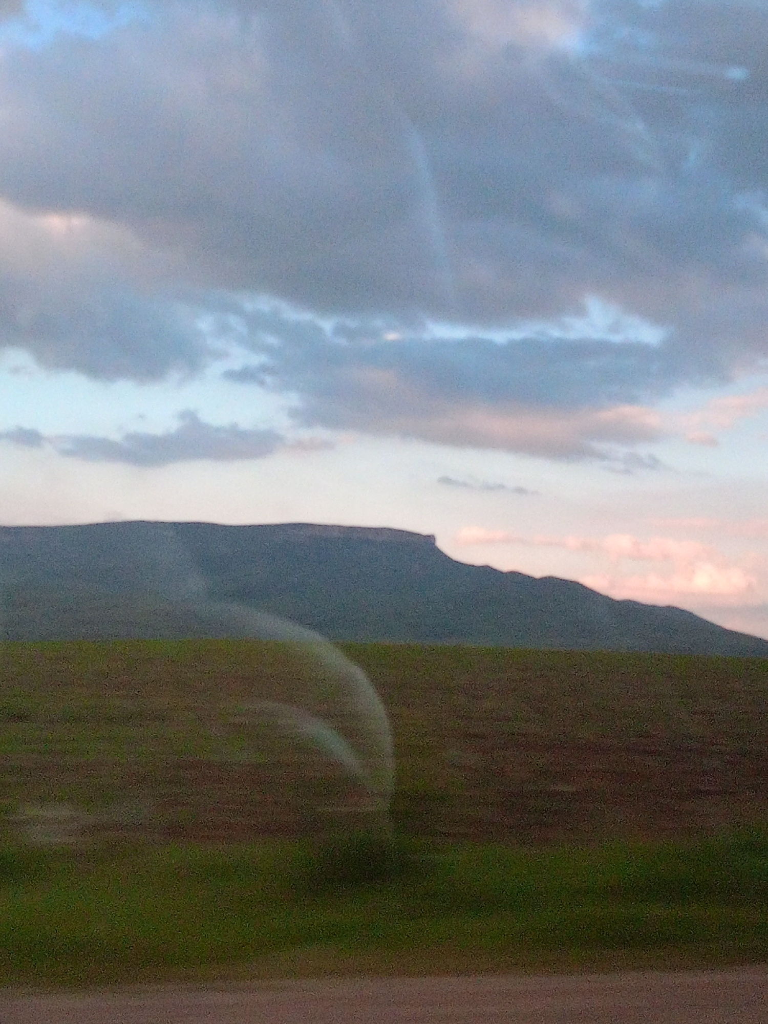 The image depicts a rural landscape with a flat-topped mountain range in the distance under a sky filled with clouds. The foreground consists of a field with green grass and brown soil, and a water sprinkler arm is visible creating a curved arc of mist over the field. The lighting suggests it is either early morning or late evening. There are no people or man-made structures immediately apparent in the scene, though the presence of a sprinkler suggests agricultural activity. This scene is consistent with the rural environment surrounding Storozhevaya, Russia, where such landscapes are common.
