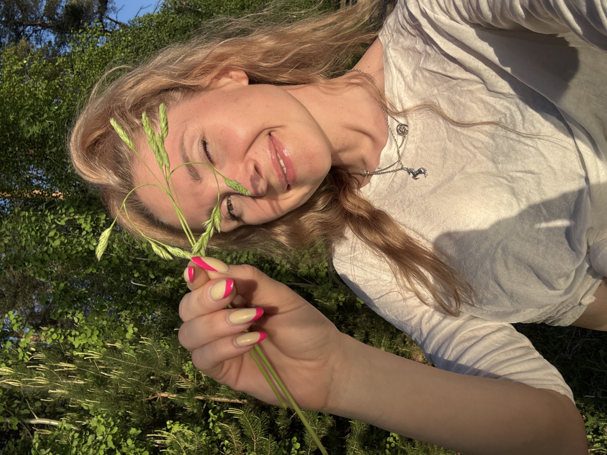 A young woman with blond hair and pink-tipped nails lies in a grassy area. She is wearing a white, long-sleeved shirt and a silver necklace with a horse pendant. She holds a sprig of green grass near her face and smiles. The background features trees and a blue sky. The location is Noviy, Russia.