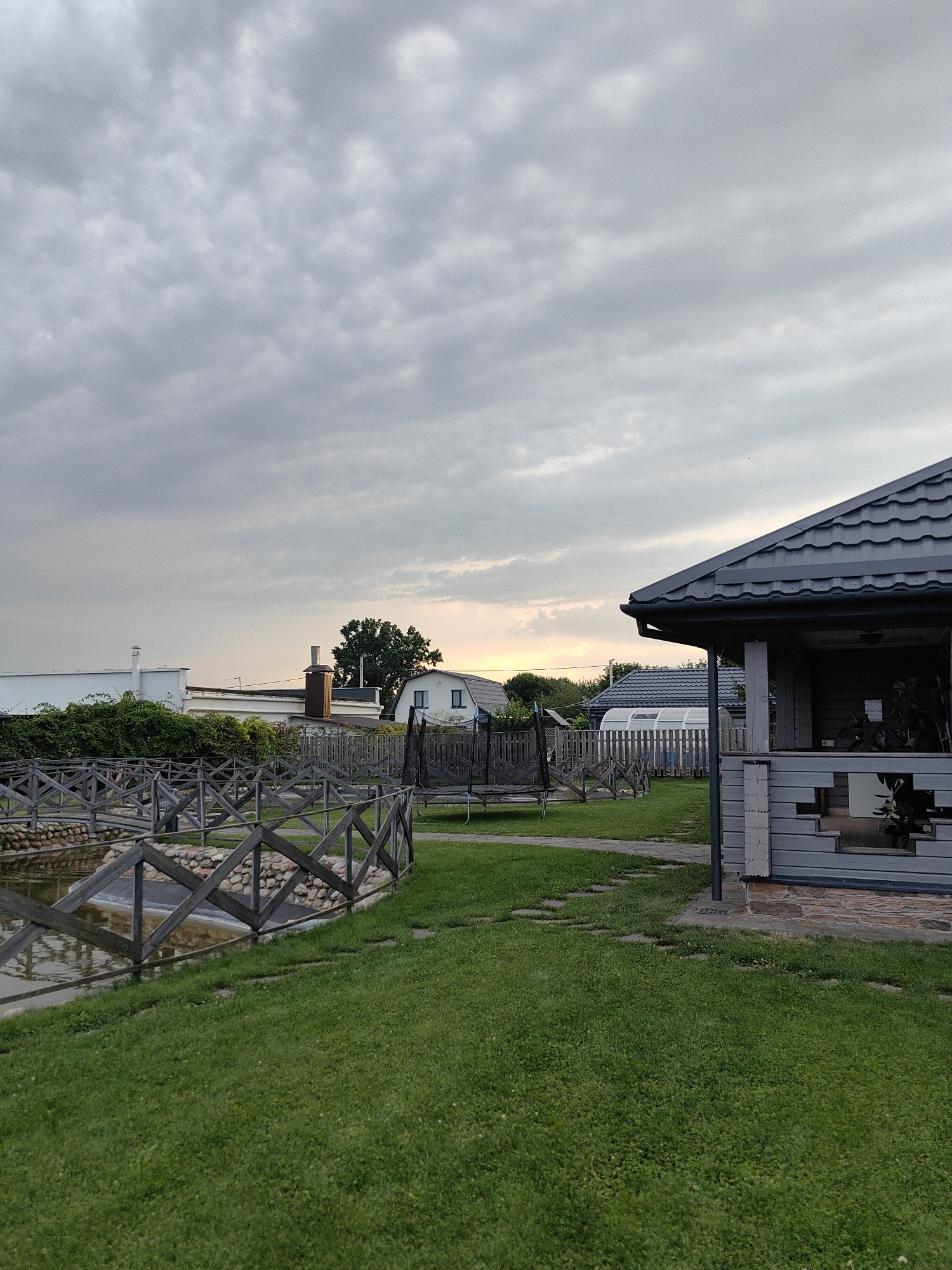 The image depicts a backyard scene in the vicinity of Фаніпальскі сельскі Савет, Belarus, under a cloudy sky. In the foreground, a lush green lawn is visible, with a winding path of stepping stones leading towards a gray gazebo on the right. The gazebo has a dark gray, tiled roof and gray wooden siding. To the left of the lawn is a body of water enclosed by a rustic wooden fence with a diamond-shaped pattern. The water appears calm, with some stones lining its edge. In the middle ground, a trampoline is set up on the grass, and beyond that, various houses and structures are visible, including a white building with a chimney, a larger house with a dark roof, and a greenhouse-like structure with a white frame and translucent panels. A large tree with dark green foliage stands behind some of the houses. The overall atmosphere is tranquil, suggesting a residential setting in a rural or suburban area. There are no people or animals present in the image.