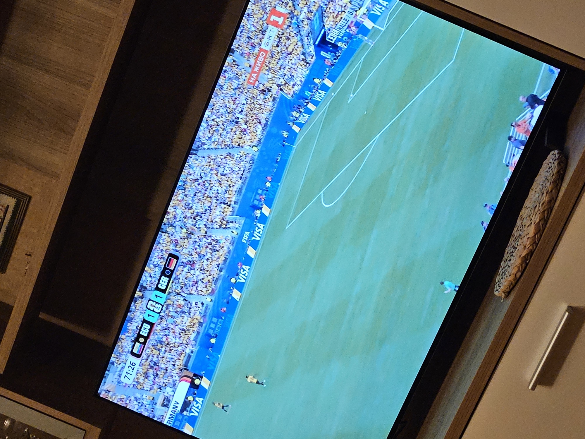 The image shows a television screen displaying a soccer match. The screen is angled, and the view is from a slight distance. The game appears to be in progress, with players on the green field. A large crowd is visible in the stands, filling the stadium. The score is displayed on the screen as 1-1, with the time at 71:26. Text overlays indicate team abbreviations, "ECU" and "GER", and the word "RMANY". Sponsor logos for "VISA" and "FIFA" are also visible along the edge of the field. A banner with the Russian text "НА ЖИВО" is also present. The lighting suggests it could be evening or an indoor stadium setting. The overall scene captures the intensity and excitement of a live sporting event.