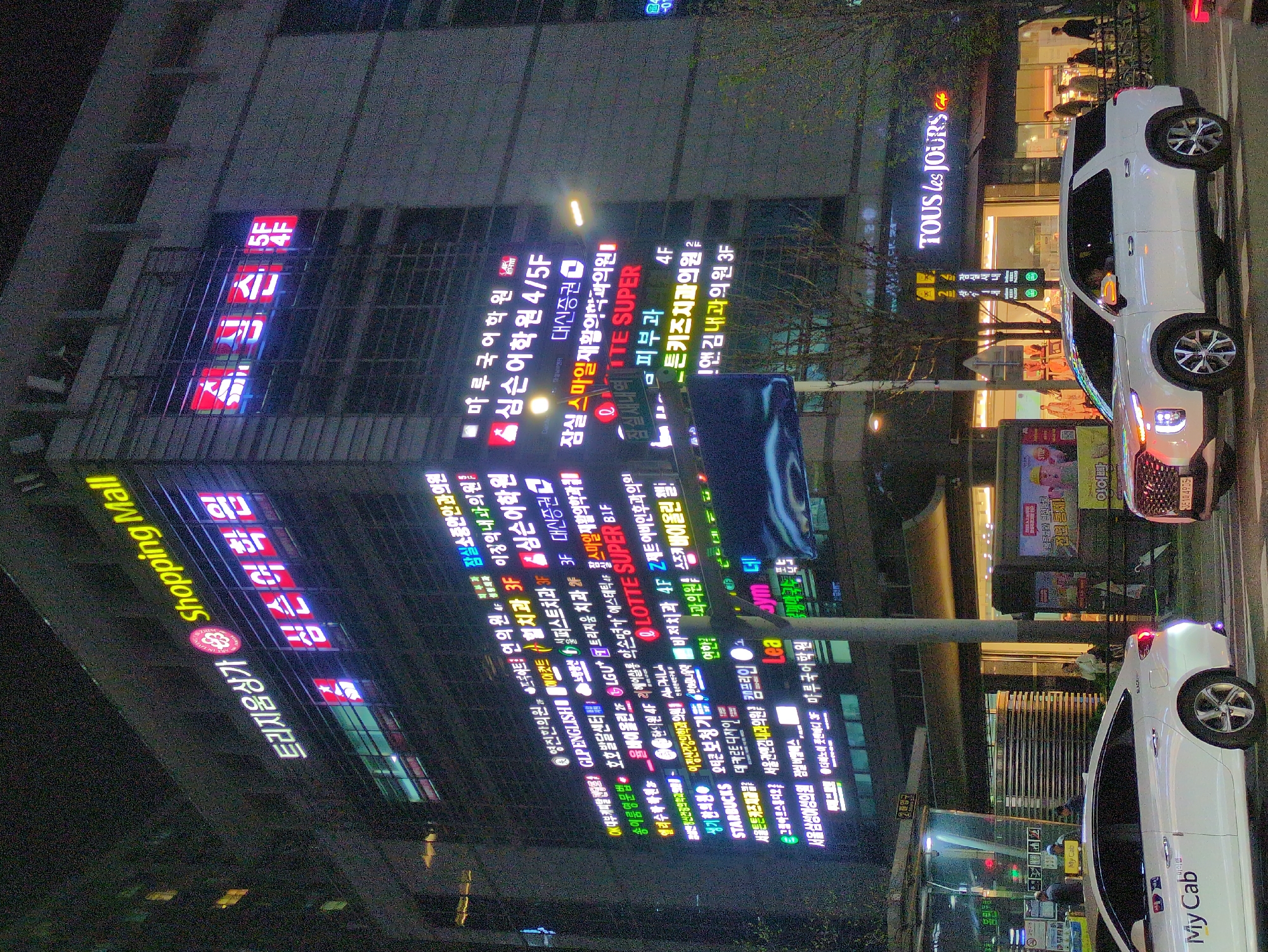 This is a night scene from what appears to be a shopping district in Seoul, South Korea. In the foreground, two white Hyundai vehicles, one a taxi ("My Cab"), are parked or moving along the street. Numerous illuminated business signs adorn a tall building, displaying various services such as language schools, medical clinics (dentists, dermatologists, internal medicine), and other businesses like Starbucks and Tous Les Jours. The signs are predominantly in Korean, but some English translations are visible. The building is identified as "Trijium Shopping Mall". Pedestrians can be seen on the sidewalk near the Tous Les Jours bakery. The street has traffic lights, and overall, the environment is brightly lit with artificial light.