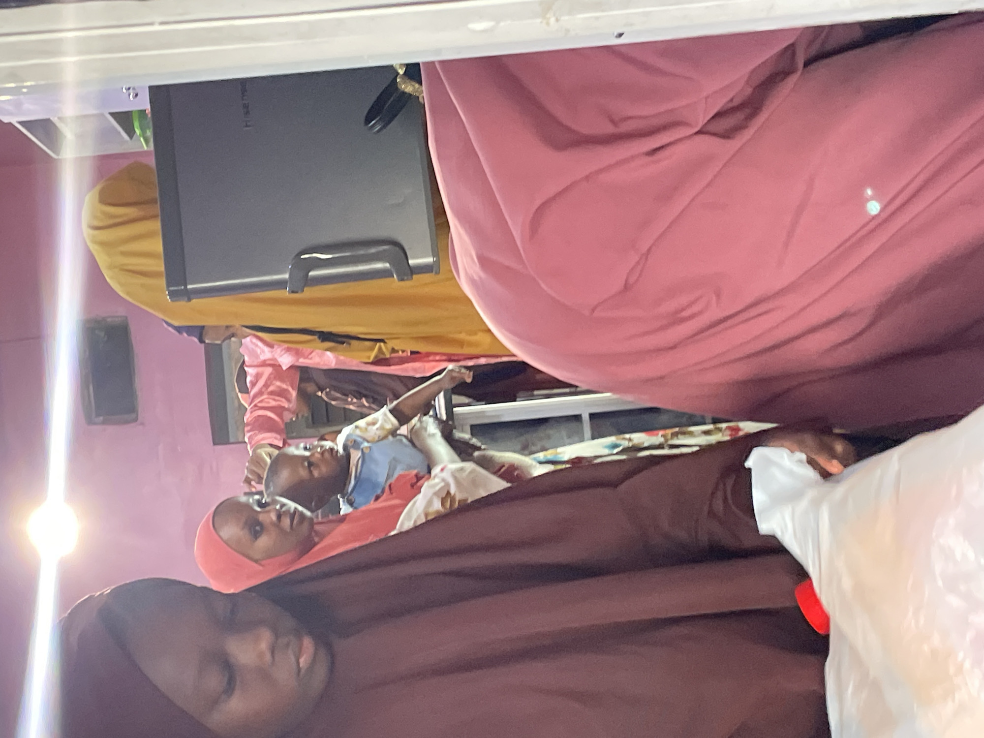 In an indoor setting, possibly a market or store in Jos, Nigeria, a group of individuals, primarily women and children, are gathered. The lighting appears to be artificial, with a bright light source visible overhead.
In the foreground on the left, a young person wearing a maroon hijab is looking downwards, holding a white plastic bag. To their right and slightly behind, a woman in a bright red hijab is looking to the side, holding a child. The child, wearing a light blue and white outfit, is being held up and appears to be looking towards the front.
Further into the background, a person in a bright yellow hijab is visible. Part of a dark electronic device, possibly a refrigerator or display unit, is on the right side of the frame. Another person in a pink garment is positioned to the right, obscuring much of the background. The walls of the room are painted a light pink color. The overall impression is one of a crowded, indoor environment.