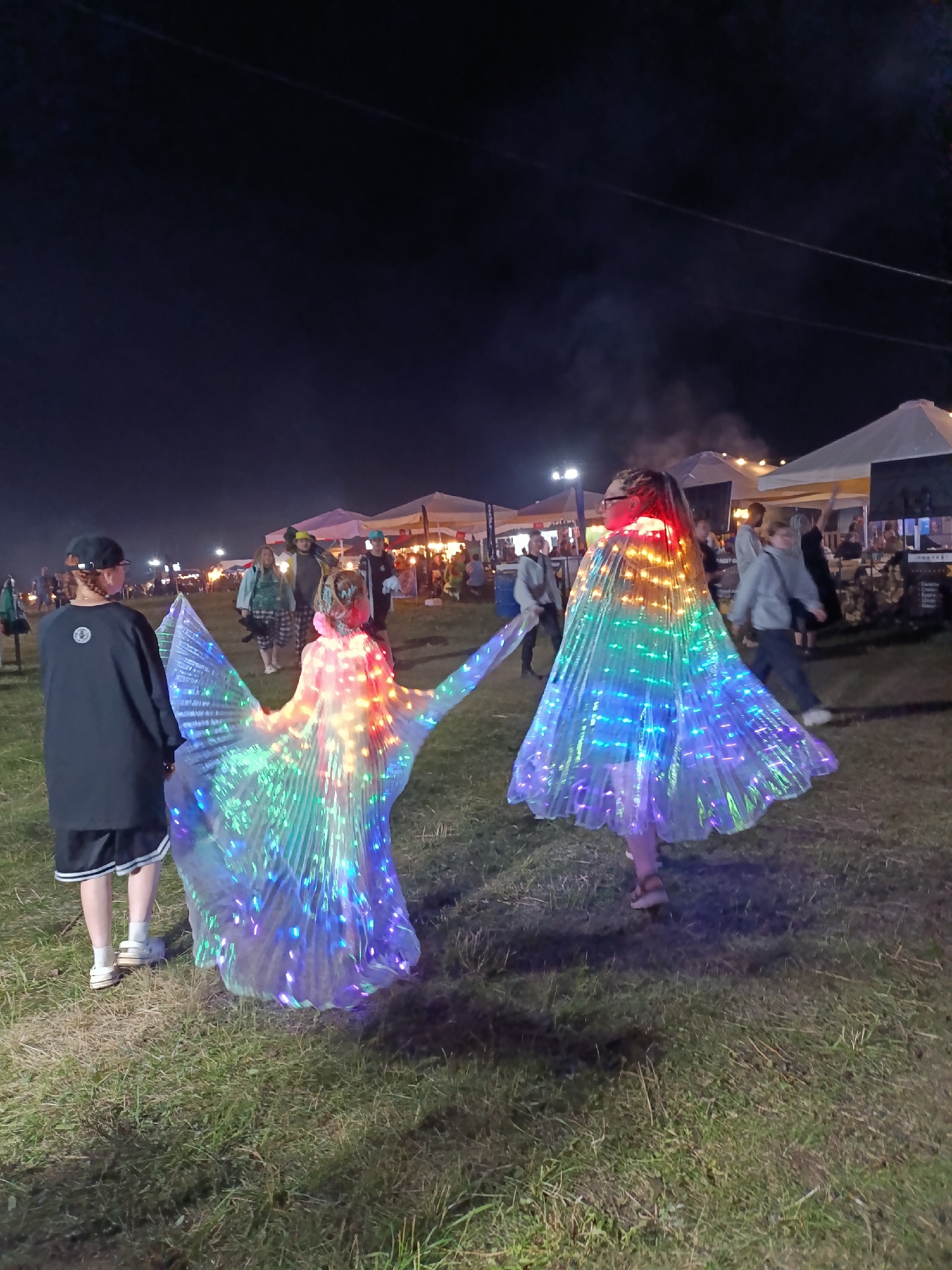 Two individuals wearing large, iridescent, LED-lit capes stand in a grassy field at night, facing away from the camera. The capes are circular and extend outwards, creating a dramatic silhouette. The person on the left has dark hair pulled back and is wearing a black t-shirt and shorts. The person on the right has braided hair and is wearing glasses. In the background, there are tents, lights, and other people, suggesting an outdoor event or festival. The scene is illuminated by artificial lights and the glow from the capes. The location is identified as Lagunovo, Russia.