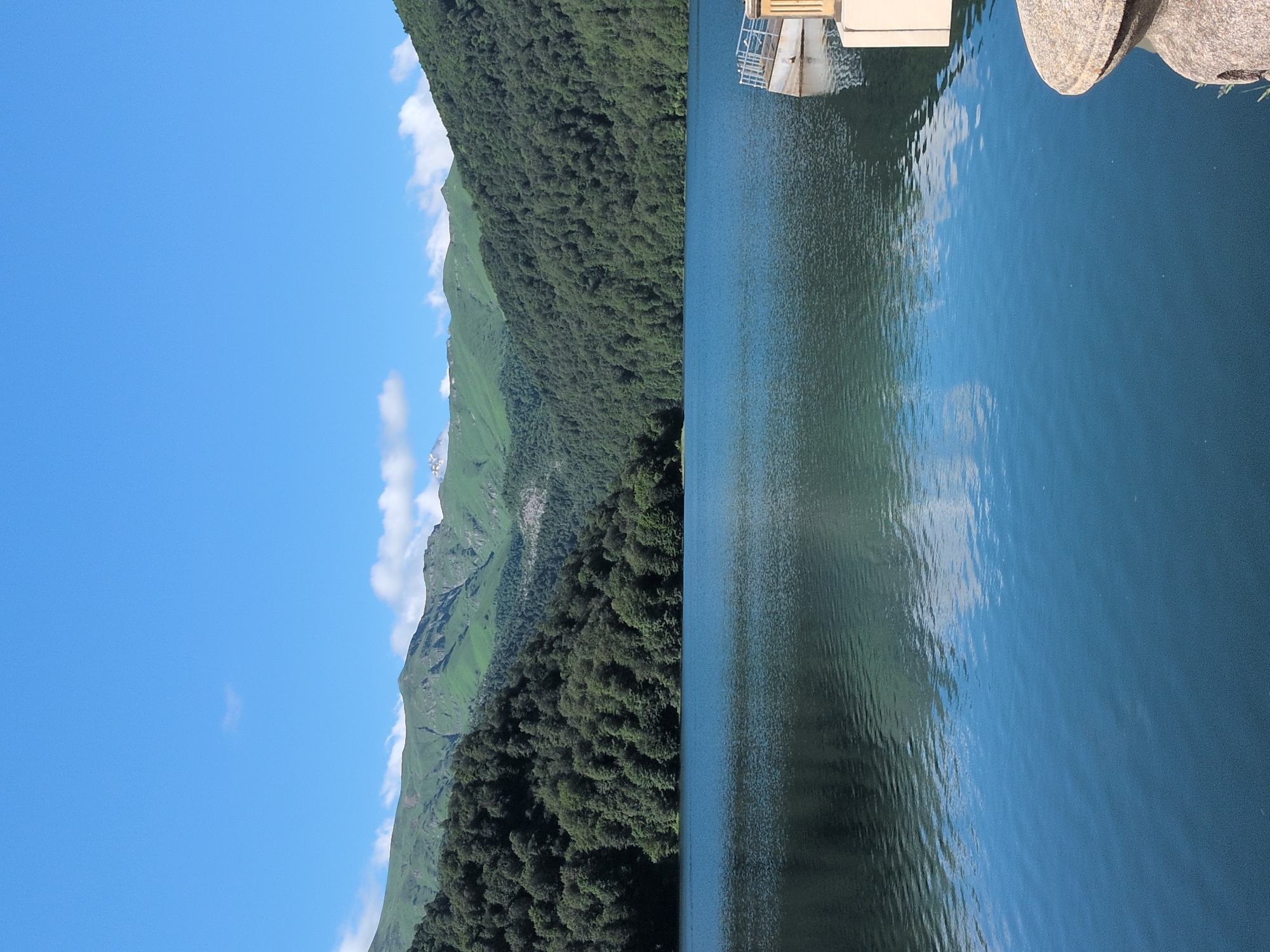 A wide shot captures a serene lake reflecting lush green mountains under a clear blue sky. The mountains, densely covered with trees, rise dramatically from the lake's edge, with some higher peaks showing hints of ruggedness. A small, white structure with railings is partially visible on the right side of the lake, near the shore. The water is calm, with gentle ripples creating a mirrored effect of the surrounding landscape. The foreground shows a textured, stone element, possibly part of a bench or monument. The image depicts a natural landscape in Toğanalı, Azerbaijan, characterized by its pristine natural beauty.