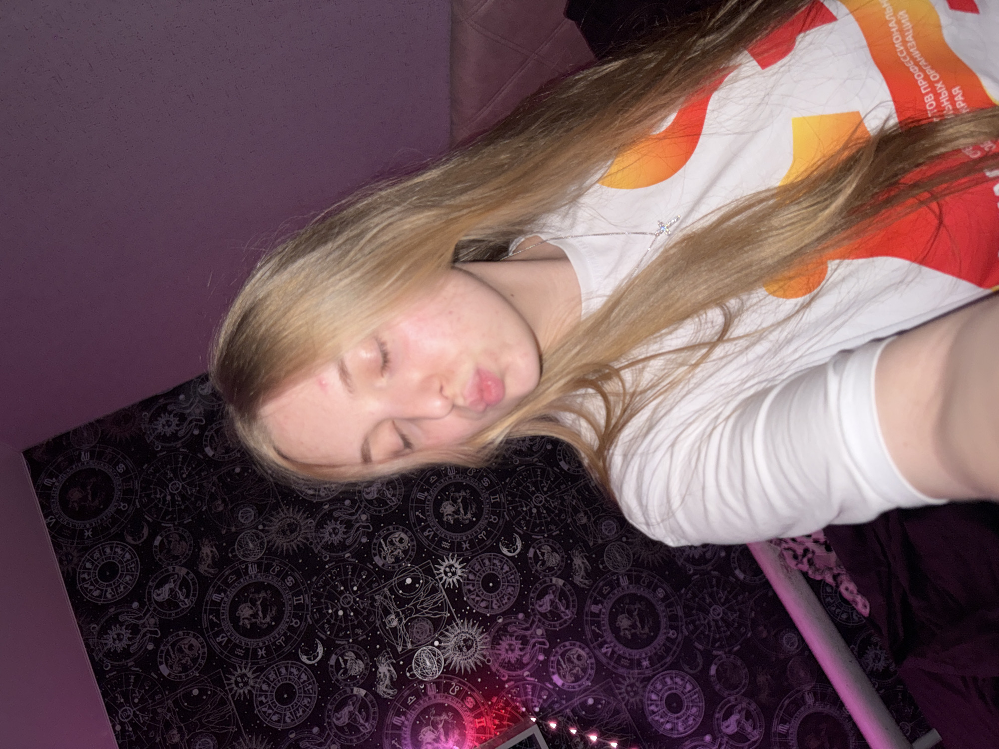 In a dimly lit room, a young woman with long blonde hair is taking a selfie. She is wearing a white t-shirt with colorful graphic print and a silver cross necklace. Her eyes are closed and her lips are puckered as if she is about to kiss. The background features a dark tapestry with astrological symbols and a pink LED light strip. A bed with a dark comforter is visible to the right of the woman. The scene appears to be set in a bedroom in an unspecified location in Russia.