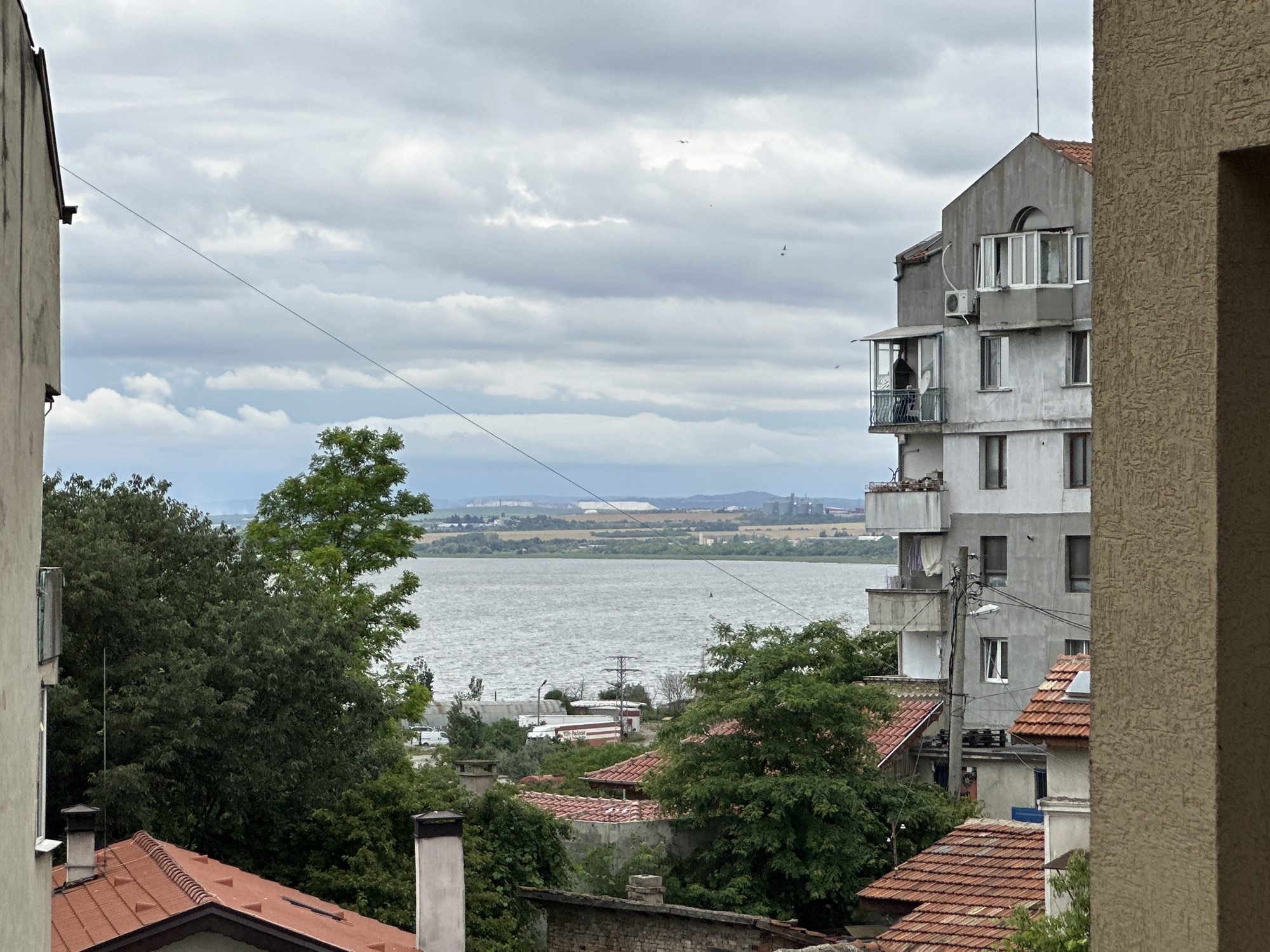 The image captures a view from a high vantage point in the city of Varna, Bulgaria, likely from a residential building. The foreground is dominated by the side of a tan, textured building on the right and a section of a lighter-colored building on the left, both partially obscuring the scene. In between, a cluster of red-tiled roofs of smaller buildings and dense green trees are visible.
Further back, a large, multi-story apartment building with a grey facade and numerous windows stands out. Several balconies are visible, one of which has a person standing on it, looking out. An air conditioning unit is mounted on the building's exterior.
Beyond the buildings and trees lies a wide body of water, likely Varna Bay, its surface rippled by wind. In the distance, across the water, a hazy industrial or port area is discernible, with various structures and a lighter colored mound, possibly a salt dome or industrial waste. The sky is overcast with heavy grey clouds, suggesting a potentially stormy or humid day. A thin wire runs diagonally across the upper portion of the image, a common sight in urban areas. A few small birds can be seen flying in the distance.