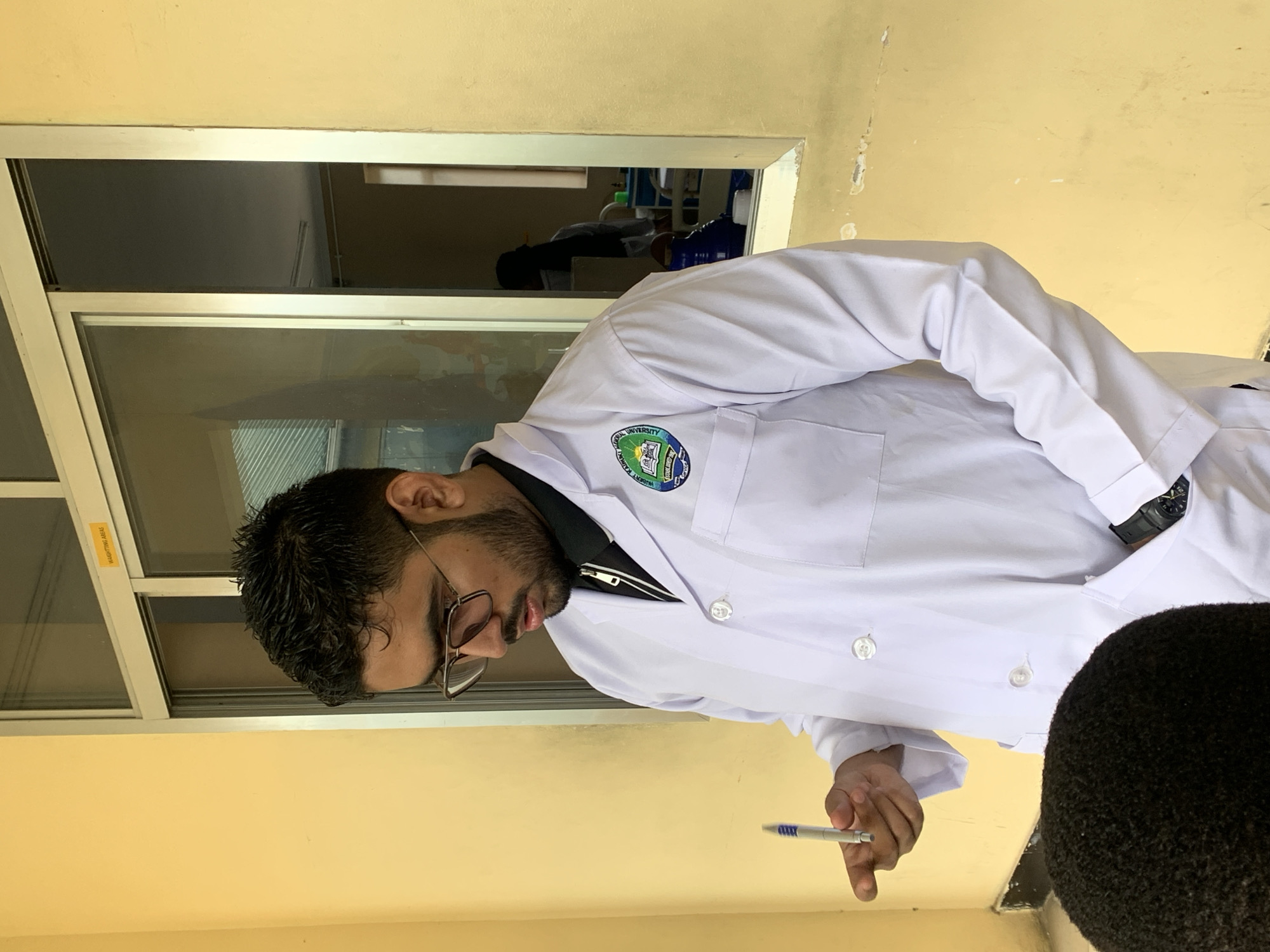 A person wearing a white lab coat stands indoors, holding a pen in their right hand. The person is facing left, looking down at the pen. They have short, dark hair, glasses, and a beard. The lab coat has a patch on the upper left chest area. The background features a window with a view into another room. The interior walls are yellow. Based on the logo on the lab coat the picture was taken at Hubert Kairuki Memorial University in Dar es Salaam, Tanzania.