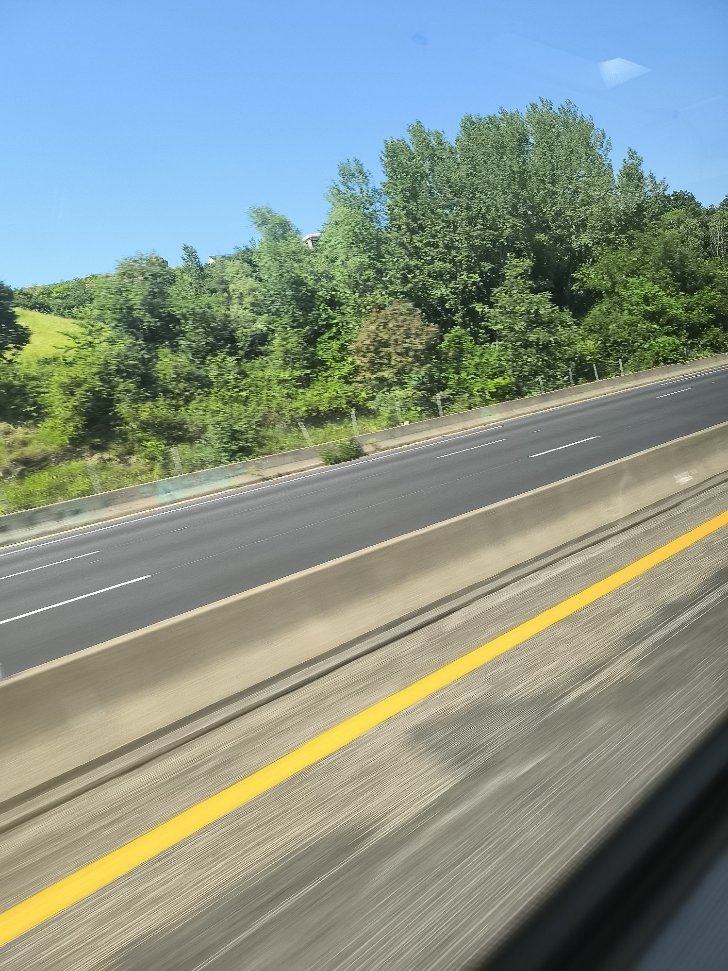 The image, likely taken from a moving vehicle on a highway in L'Horme, France, shows a multi-lane roadway with a concrete barrier and a solid yellow line separating the shoulder from the traffic lanes. Lush green trees and vegetation line the opposite side of the highway. The sky is clear and blue. The perspective suggests motion. There are no visible people or other vehicles.