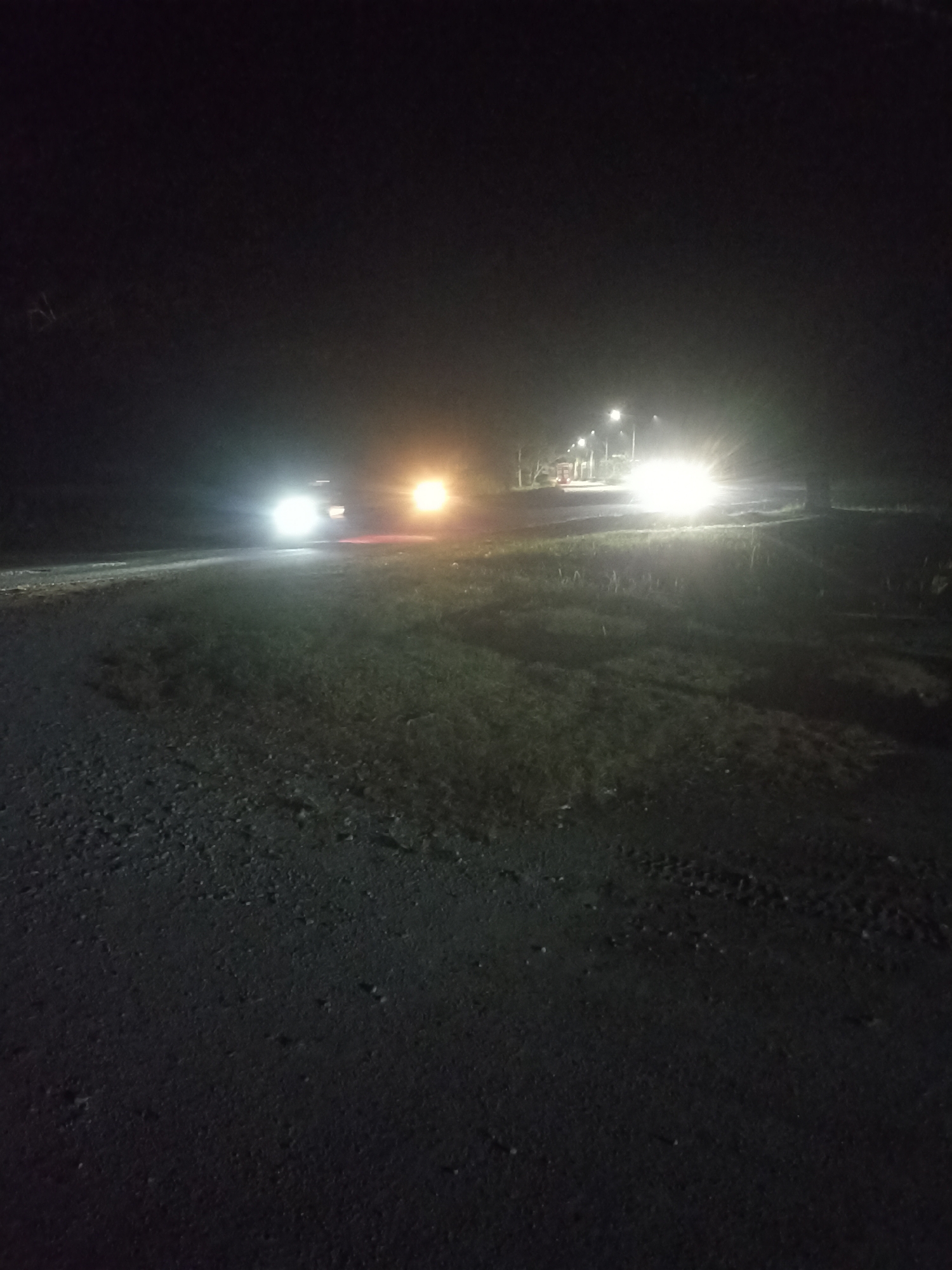 The image shows a rural road at night in Kampung Tengah, Indonesia. The road is unpaved and appears to be made of gravel and dirt, with some grassy areas on the sides. Two vehicles are visible on the road, both with their headlights on, illuminating the surroundings. The vehicle on the left has its headlights on bright, casting a strong beam. The vehicle on the right has its headlights on as well, and there appears to be a warm-toned light, possibly from a street lamp or another vehicle, further down the road. The scene is dark, with the headlights of the vehicles being the primary source of light. The environment suggests a remote or less developed area, typical of a village setting. There are no people visible in the image. The overall impression is one of nighttime travel on a country road.