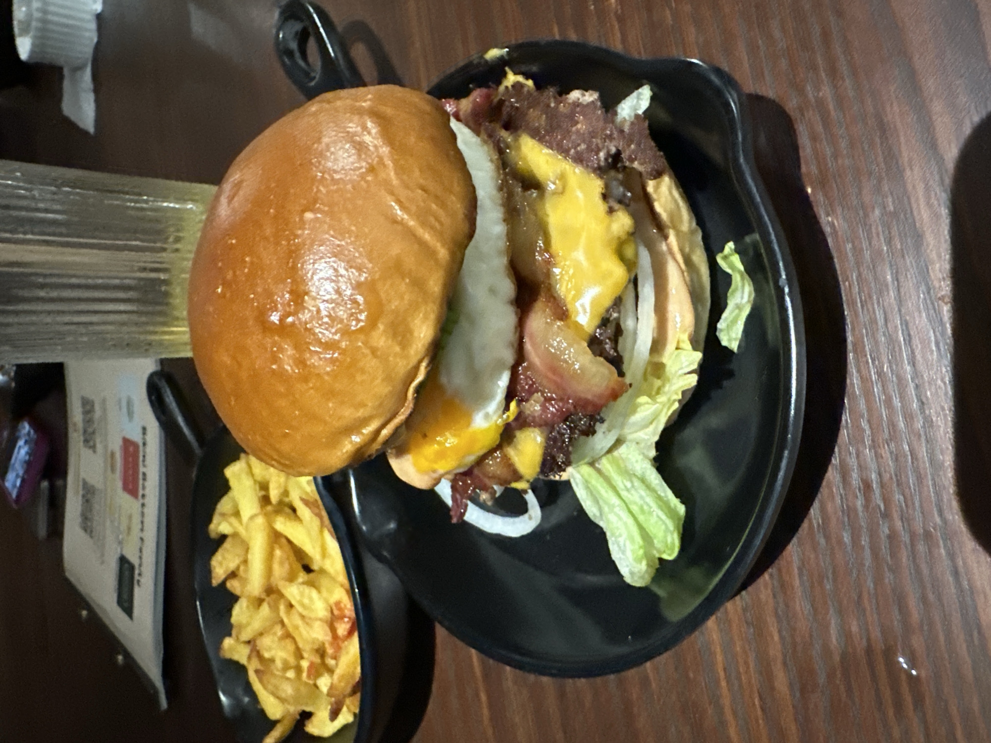 On a dark wooden surface is a black plate containing a hamburger and fries. The hamburger features a glazed bun, lettuce, white onion, a possible fried egg, cheese, bacon, and ground beef. Next to the burger is a separate small black dish with fried potatoes. Nearby, on the left side of the frame, is a clear glass bottle with a white top and a menu with visible text and QR codes. The setting suggests a restaurant or eating establishment. The image location is likely Đà Nẵng, Vietnam.