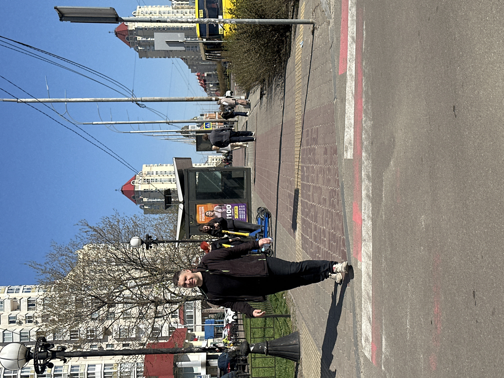The image shows a man standing on a sidewalk in Kyiv, Ukraine, with a street to his right. He's facing the camera, wearing a black jacket, black pants, and white sneakers. The sidewalk is paved with light brown bricks and bordered by a red and white painted stripe. In the background, there are several multi-story apartment buildings, some with red roofs. Utility poles with numerous wires are visible in the upper left. A bus stop with advertisements stands behind the man. People are seen standing and walking on the sidewalk in the distance. The weather appears sunny, with a clear blue sky overhead.