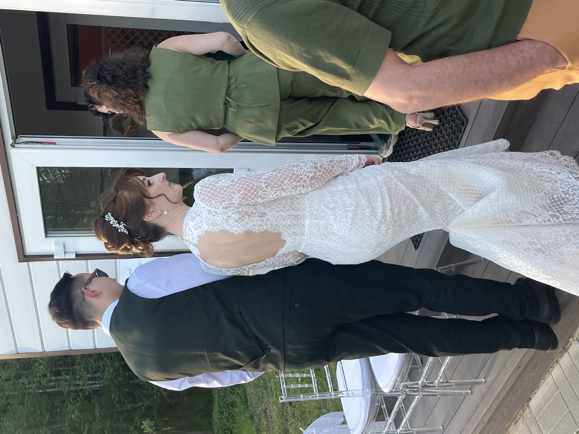 The image shows a wedding scene outdoors. A bride in a white, lace wedding dress with an open back stands with a groom wearing a black vest, white shirt, and black pants. They are positioned near what appears to be a doorway and white plastic chairs. Another person, dressed in green, stands near the doorway. Another individual, wearing what seems to be a green or mustard-colored shirt, stands to the side. The location may be Алопово, Russia, but there are no contextual clues to confirm. The environment consists of wood flooring or decking and green foliage.