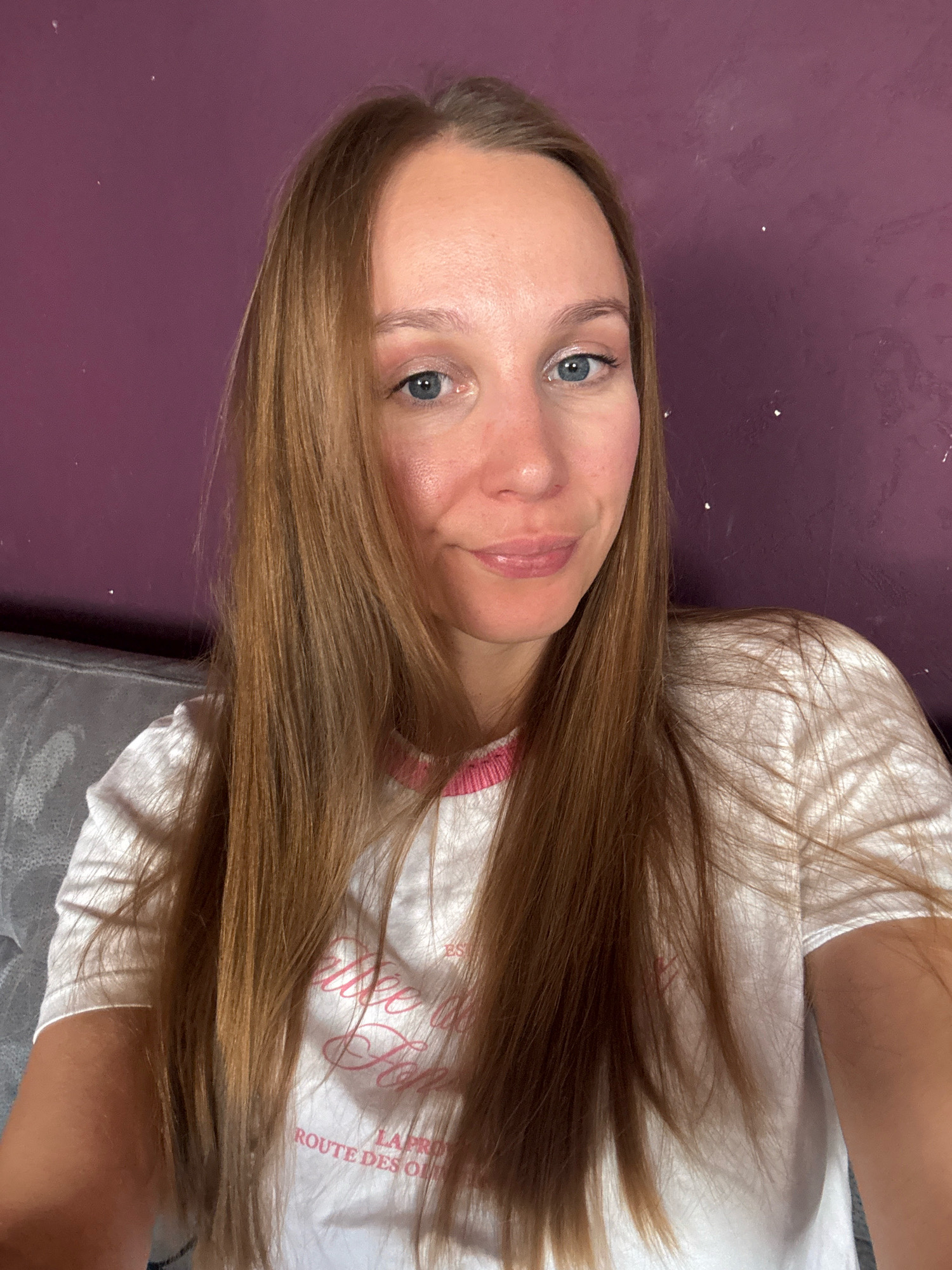 A young woman with long, light brown hair and blue eyes smiles at the camera. She is wearing a white t-shirt with pink trim around the neckline. The background is a textured purple wall. The image appears to be a selfie, taken indoors in a residential setting. The woman is in the foreground, with the wall behind her. There is a hint of a gray couch to the left of the frame. The image does not contain enough information to determine the location in the city of Überherrn, Germany.