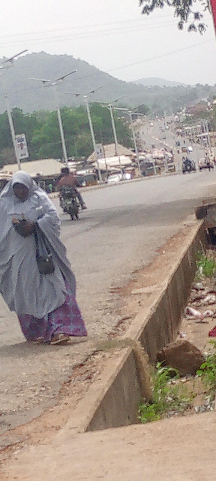 This image captures an everyday street scene in Adankolo, Nigeria, featuring people, vehicles, and distant hills under what appears to be an overcast sky.
In the foreground, slightly to the left, a woman walks along the side of a paved road. She is wearing a light-colored, full-length garment (possibly an abaya or hijab) that covers her head and body, with a visible patterned purple and blue skirt beneath. A dark, small shoulder bag is visible, and she seems to be looking down at an object in her hands.
Further into the middle ground, a man rides a motorcycle on the road, seen from behind. He appears to be bare-chested. The road continues into the distance, populated by other blurry vehicles and figures, indicating traffic. Along the road, there are structures that resemble market stalls or shops. Several modern street lights, possibly with solar panels, are visible along the roadside, extending towards the background.
To the right of the walking woman, a concrete curb or drainage ditch runs parallel to the road, containing some green vegetation and indistinct debris. In the far background, hazy green hills or mountains dominate the skyline, suggesting the topographical features of the region. The overall scene depicts typical daily activities in a bustling urban or semi-urban environment. No specific text is clearly legible on any signs or objects within the frame.