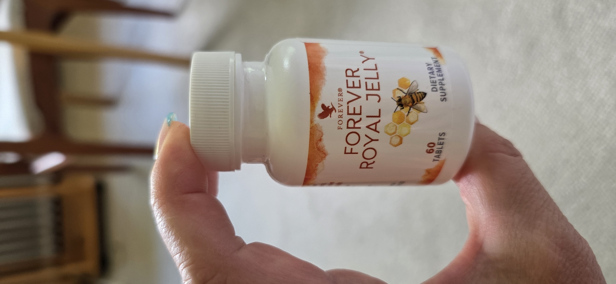 A hand holds a white bottle of Forever Royal Jelly dietary supplement. The bottle has orange and yellow honeycomb patterns and a depiction of a bee and the Forever Living Products logo. The text "FOREVER ROYAL JELLY" is prominent on the label, along with "60 TABLETS" and "DIETARY SUPPLEMENT." The background is blurred, showing the legs and seat of wooden chairs, suggesting an indoor setting, possibly in Maasbüll, Germany.