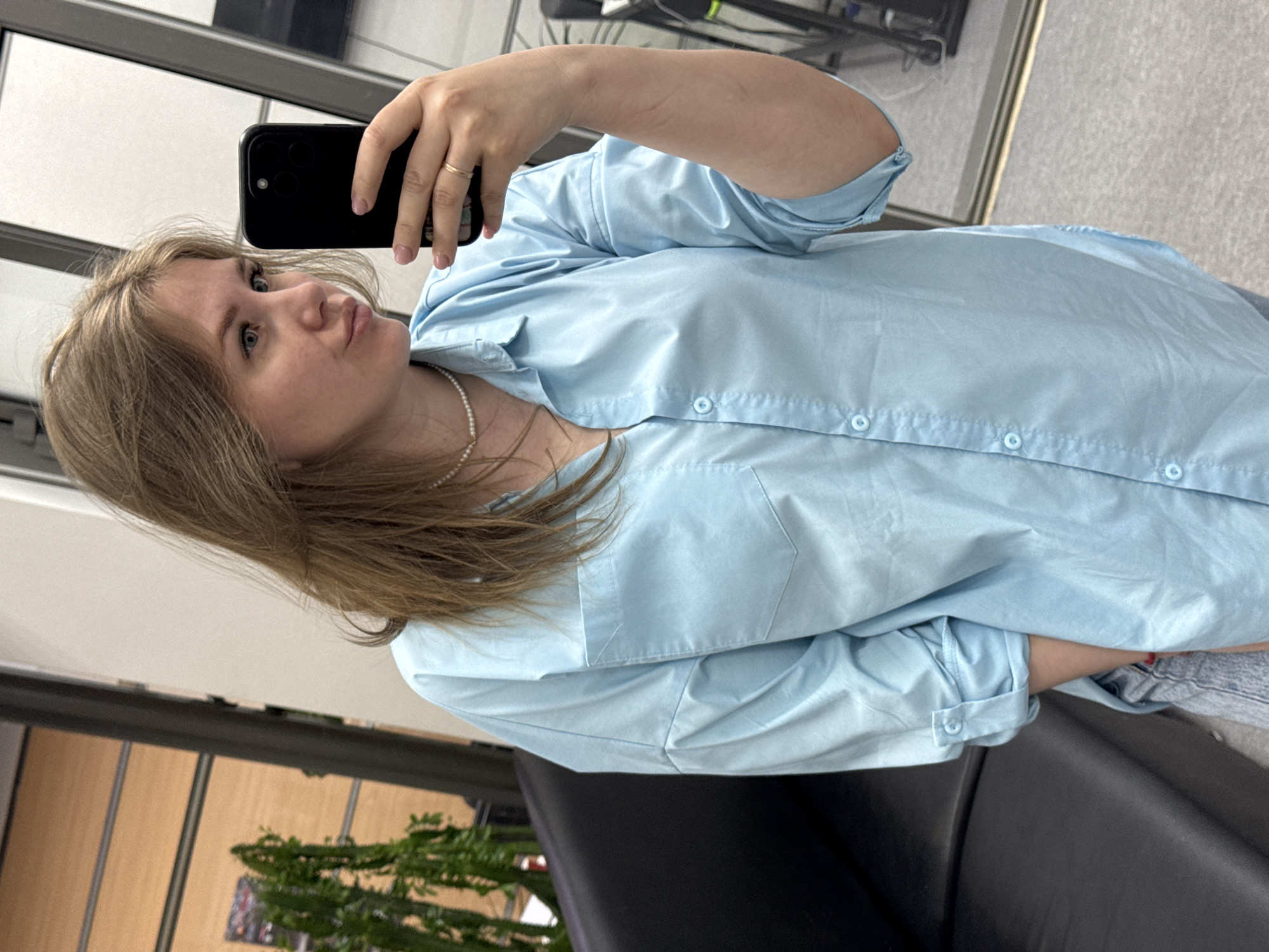A fair-skinned woman is taking a selfie using a black smartphone. She is wearing a light blue button-down shirt and a pearl necklace. A ring is visible on the ring finger of her left hand. She is positioned in front of a window and a black chair. Behind the chair, there is an indoor plant. The image was taken in Perm, Russia.