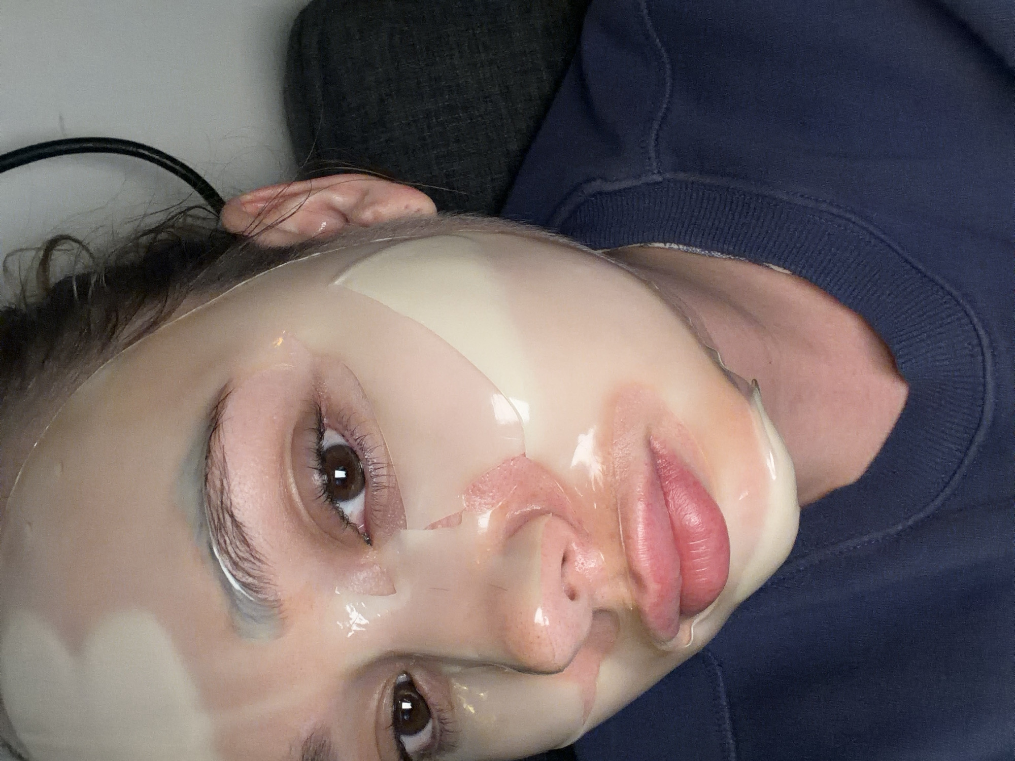 A close-up, tilted shot shows a person wearing a sheet mask. The person's face is turned towards the viewer, with their eyes open. The mask is translucent and covers most of the face. A black cable is visible behind the ear. The person is wearing a blue sweatshirt. The location is not discernible from the image and cannot be determined to be in Ramat Gan, Israel.