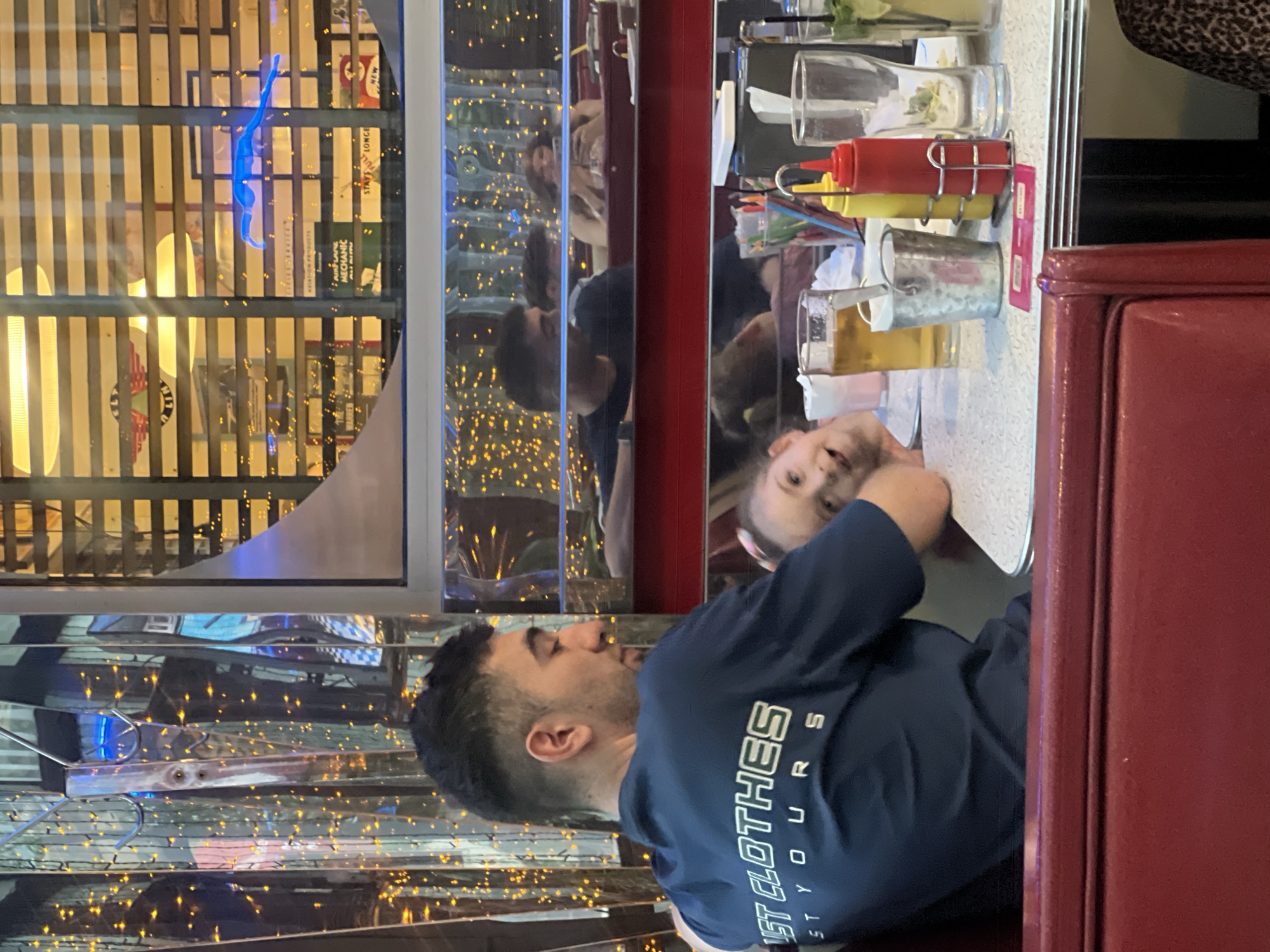 The image captures a scene inside a restaurant with retro-style decor. A man wearing a navy blue t-shirt with white lettering is seated at a table, with his back to the viewer. Across from him, a young girl looks towards the camera. The table has condiments (ketchup and mustard bottles) and drinks on it. Through a window beside them, a lit-up display can be seen, featuring a blue neon dinosaur. The restaurant appears to be decorated with numerous small lights, possibly giving the scene a festive or vintage atmosphere.
