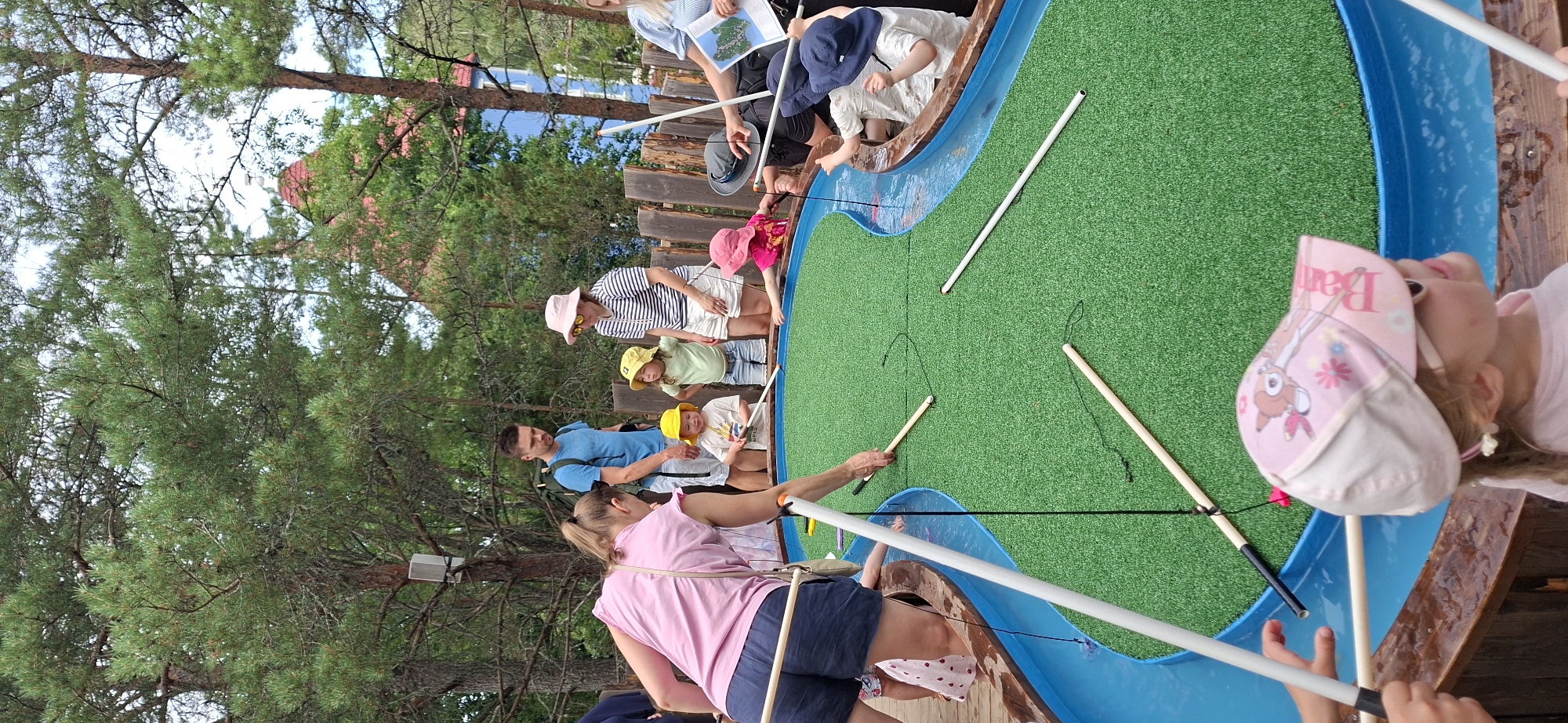 The image depicts a group of adults and children engaged in an activity, likely a miniature golf course designed with a water feature. The setting appears to be outdoors, with lush green trees and a hint of a red-roofed building in the background, suggesting a park or recreational area. The primary focus is on a blue, shallow water channel with a bright green, artificial turf surface. Several individuals are interacting with long, white poles, some of which are attached to strings. A woman in a pink shirt and blue shorts is positioned on the left, leaning over the water with a pole in her hand. Other adults and children, many wearing hats, are gathered around the course, observing and participating. The children are actively engaged, with some holding poles and others looking towards the water. The overall atmosphere is one of casual recreation and family fun. The building in the background, with its distinctive red roof and blue facade, is reminiscent of architecture found in Moominworld in Naantali, Finland, suggesting this scene may be taking place within that amusement park.
