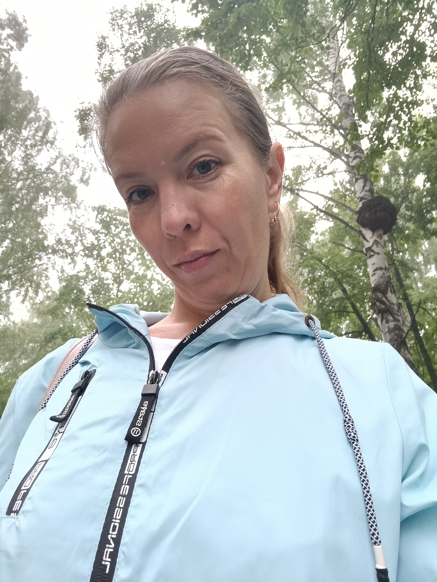 A low-angle outdoor selfie captures a woman, likely in her 30s or 40s, looking directly at the camera. She has light-colored hair, pulled back, revealing blue eyes and a neutral, slightly contemplative expression. She is wearing a light blue hooded jacket, possibly a windbreaker, over a white t-shirt. The jacket features black zipper straps with white text; on the main zipper, the text appears to be "JAVIOI2ES3" and "SYSTEM," while "SYSTEM" is also visible on a pocket zipper. A black and white patterned drawstring hangs from the hood. A small gold hoop earring is visible in her right ear.
The setting is an overcast outdoor environment, consistent with Chaikovsky, Russia. In the background, several trees are visible, prominently including a birch tree with its distinctive white bark and black markings. A large, dark, irregular growth, possibly chaga fungus or a burl, is prominent on the birch tree's trunk. The soft, diffused lighting suggests an overcast day. The woman is stationary, posing for the photograph.