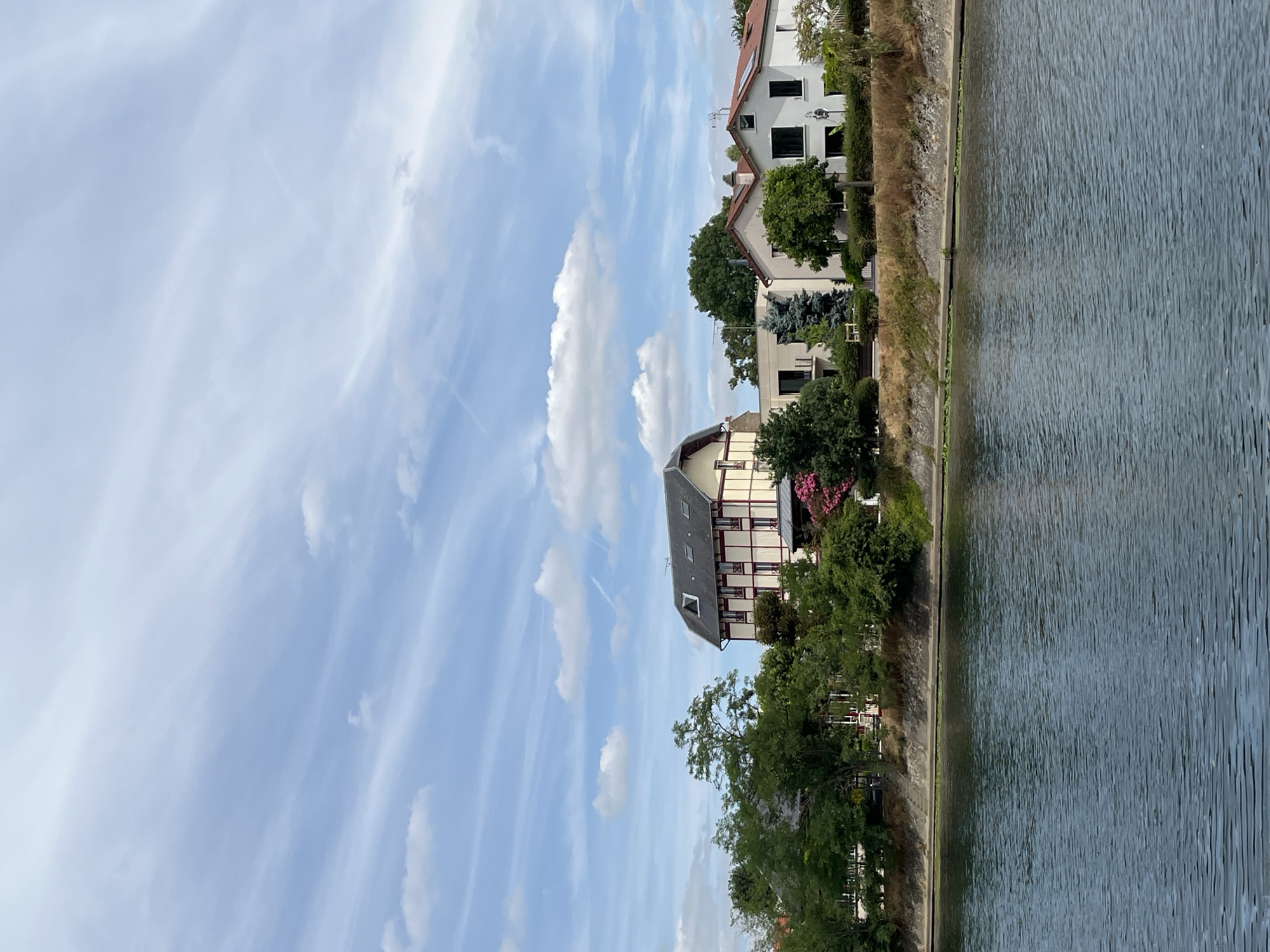 The image shows a body of water in the foreground, reflecting the sky above. The water is dark blue and appears calm, with gentle ripples on its surface. On the far bank, a line of trees and vegetation separates the water from a residential area. Several houses are visible, varying in architectural style and color. The most prominent building is a large, light-colored house with dark trim and a dark roof, featuring multiple windows. Other houses in the background are white with red tile roofs. The sky is blue with scattered white clouds, some of which are wispy and streaky, suggesting high-altitude clouds or contrails from aircraft. The scene depicts a peaceful, sunny day in a suburban or semi-rural setting, likely along a river or canal in Joinville-le-Pont, France.
