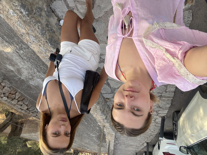 An outdoor eye-level selfie-style photograph depicts two young adult females on a stone-paved pathway in the village of Deià, Spain. The pathway slopes downwards from left to right.
The female in the foreground on the left has light skin, light brown hair pulled back, and wears a pink long-sleeved tunic with white lace trim on the sleeves and collar, featuring a V-neck with tassels, over a multi-colored bikini top. She holds the camera, angled slightly upwards towards herself and the second individual.
The second female, positioned behind and to the right, has light skin, long straight brown hair, and wears a white spaghetti-strap tank top over a blue bikini top, paired with white denim shorts and black sandals. A black digital camera with a neck strap hangs from her neck, and a black shoulder bag is visible on her left shoulder. She stands on the sloped path, looking directly towards the camera.
To the left of the subjects, a stone-paved road accommodates two parked white vehicles. To the right, a stone retaining wall with a wire fence borders a hillside featuring dry, sparse vegetation and an olive tree. The lighting suggests late afternoon or early evening.