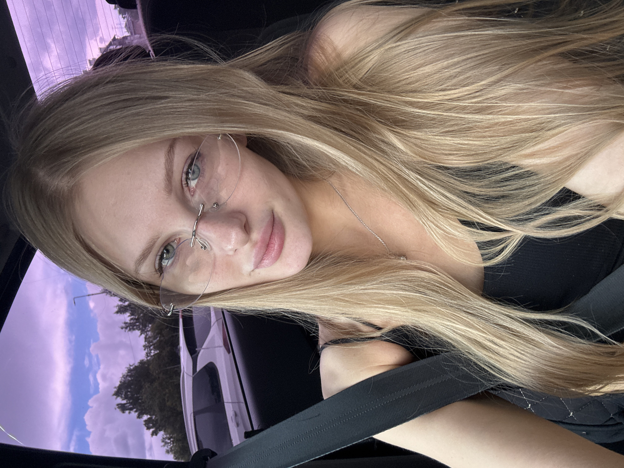A young woman with long blonde hair and blue eyes is seated in the passenger seat of a car, looking into the camera. She is wearing rimless glasses, a black tank top, and a silver necklace. Her seatbelt is fastened. The background outside the car windows shows a cloudy sky with purple hues, trees, and other vehicles. The scene appears to be set in a parking lot or on a street within the city of Perm, Russia, given the distinctive purple tint of the sky and the architectural style of the buildings visible in the distance.
