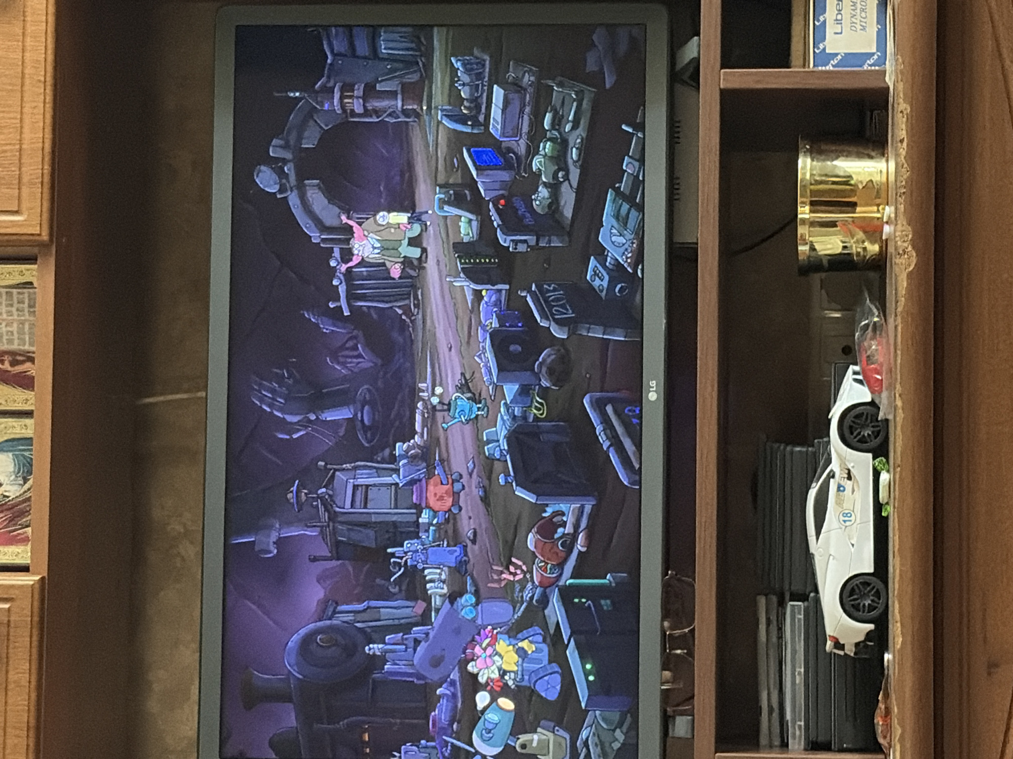 The image shows a television screen displaying a scene from a cartoon. The cartoon depicts a gritty, industrial-looking environment with a large train in the background. In the foreground, several small robot-like characters are scattered around, some of which appear to be damaged or disassembled. One larger character, a man with pink hair and a brown jacket, stands near a stone archway. Below the television, on a wooden shelf, is a white toy sports car, a stack of DVDs, and a gold-colored cylindrical object. To the right of the gold object is a blue and white box labeled "Liberty". The overall scene suggests a post-apocalyptic or dystopian setting within the cartoon.