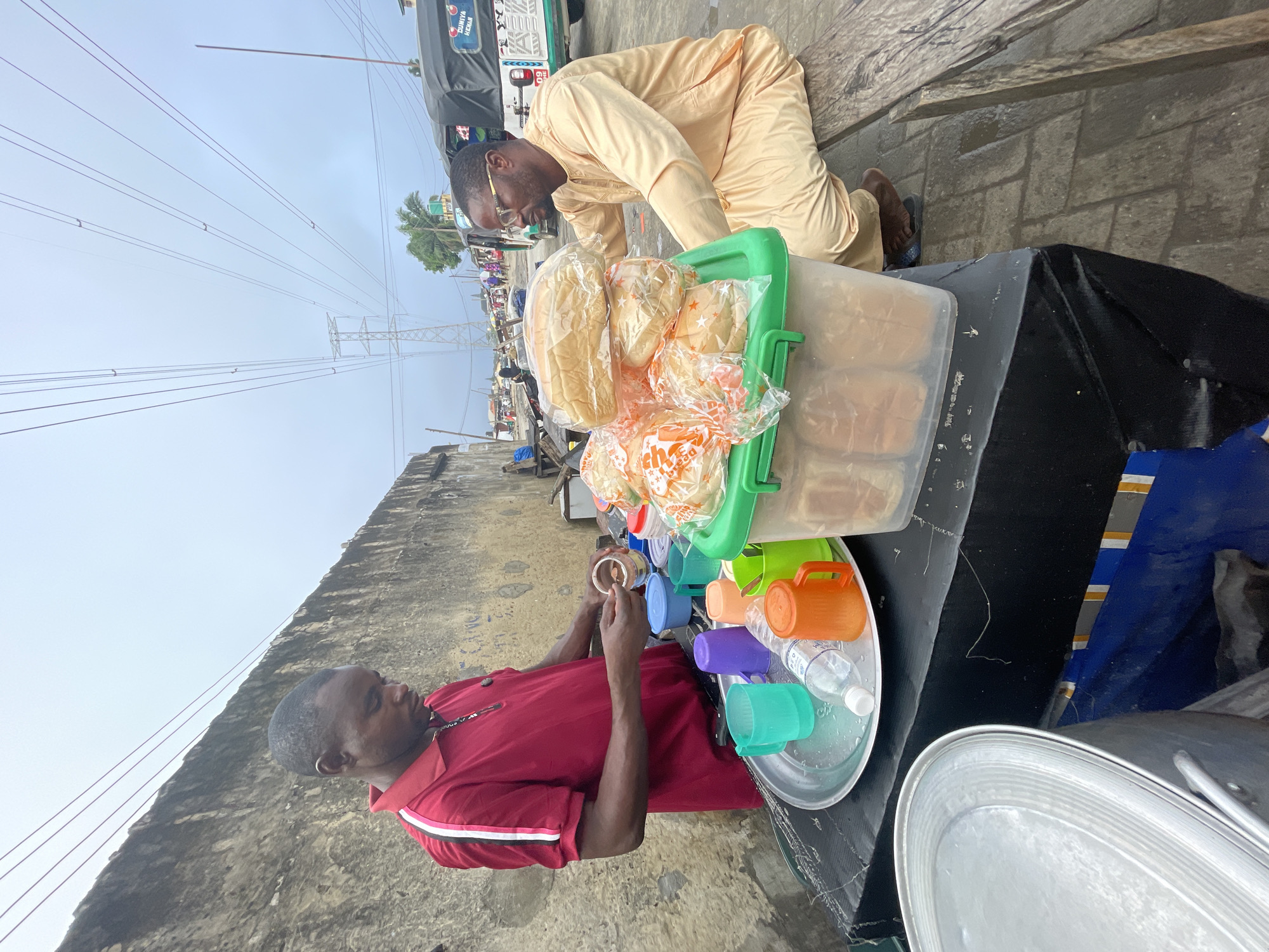 The image depicts a roadside scene in the city of Ado, Nigeria, featuring two men and a vendor stall.
In the foreground left, a large, lidded aluminum pot is partially visible. Centrally, a makeshift vendor stall with a black, fabric-draped surface supports various items. On the stall, a circular silver tray holds a clear plastic water bottle and multiple colorful plastic mugs (teal, purple, orange, green).
Standing behind the stall, to the left, a man wearing a red polo shirt with white and pink stripes on the sleeves is engaged in preparing a substance. He holds a spoon and is scooping contents from a small brown container, possibly into a cup or another vessel. His gaze is directed downwards at his task.
To the right of the red-shirted man, on the stall, rests a green-lidded clear plastic container filled with individually packaged bread rolls or buns. Several additional plastic bags containing loaves of bread are stacked on top of this container.
To the right of the stall, a second man, wearing a tan long-sleeved tunic and trousers, is seated on a wooden bench or plank, facing towards the bread display. He wears eyeglasses and is observing the items on the stall. His bare feet, clad in dark sandals, are visible.
The background features a weathered concrete or stone wall running horizontally behind the vendor's setup. Above the street, multiple electrical wires stretch across an overcast, light grey sky. A large electricity pylon is discernible in the distant haze. Further down the street, various structures, pedestrians, and a black and white motorized tricycle (Keke Napep), bearing the text "DUNIYA KENAN" and "09 HE" on its rear panel, are visible. The ground surface consists of grey interlocking paving stones, some appearing damp.