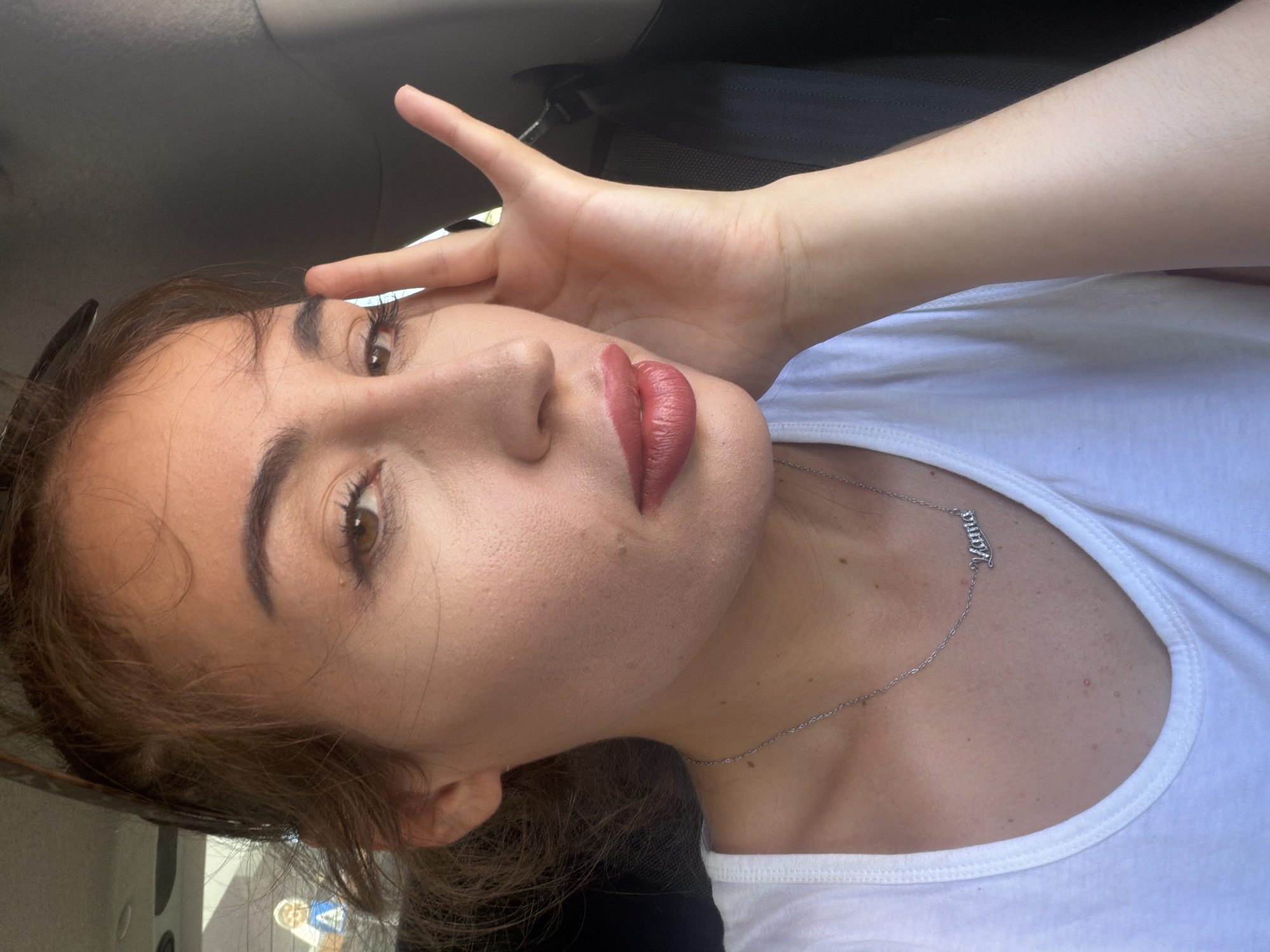 A close-up, head-and-shoulders shot captures a woman from the chest up, seated inside a vehicle. The woman, facing slightly to the right and angled toward the camera, has medium-toned skin with visible pores and freckles. Her hair is pulled back in a casual, light brown ponytail with loose strands framing her face. She is wearing a white V-neck t-shirt and a silver necklace with a pendant spelling out "Mama" in cursive. Her makeup includes foundation, mascara, and a matte, pinkish-red lipstick. Her right hand is raised to her head, with her fingers gently touching her temple.

The background is predominantly dark, suggesting the interior of a car. A sliver of bright light is visible to the left of the woman's head, hinting at an outdoor environment. The image provides no specific landmarks or details to confirm a location in Kiti, Cyprus.