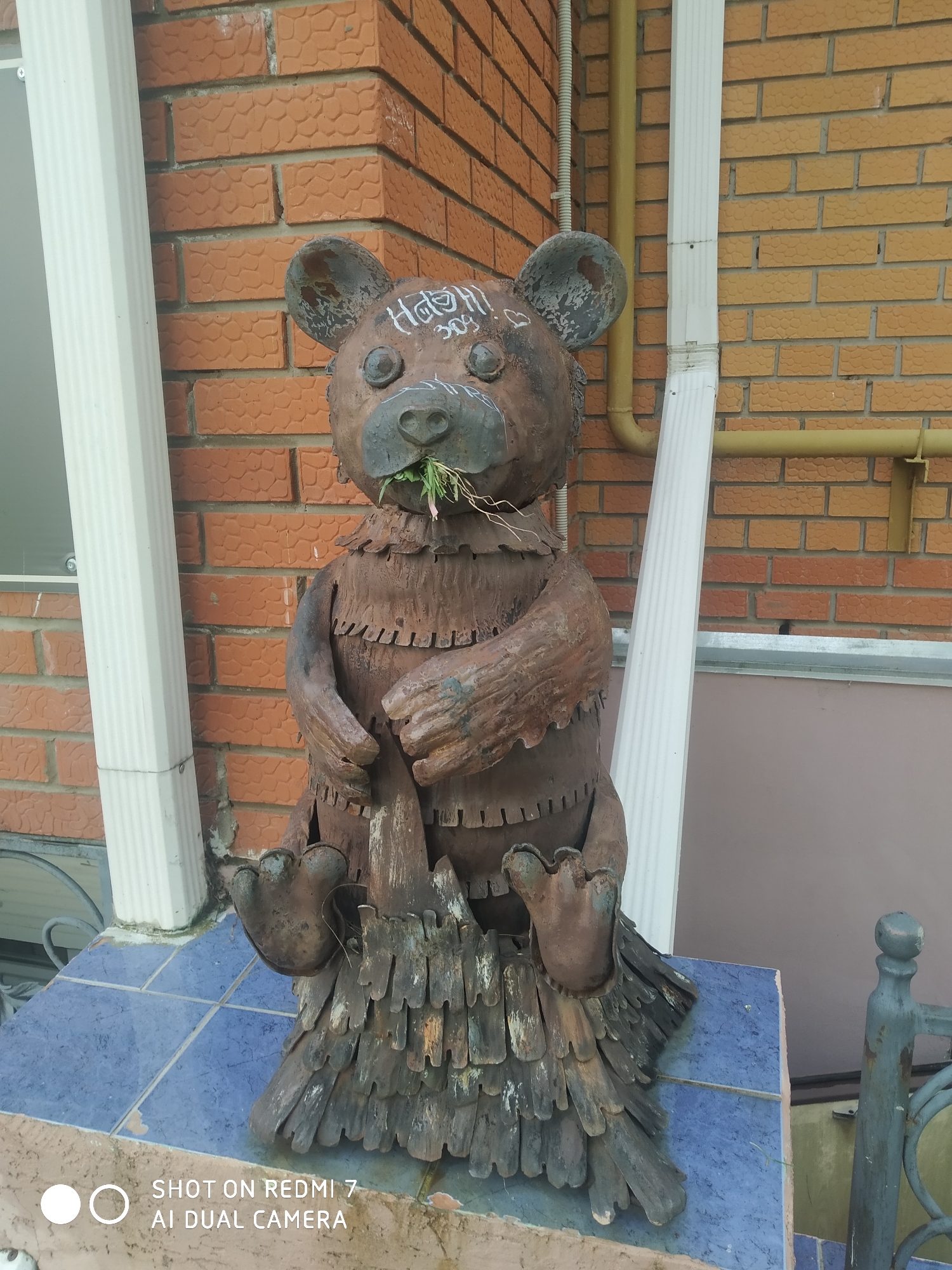 A brown, metal bear sculpture stands on a blue tiled surface outside of a brick building. The sculpture appears to be crafted from various pieces of metal. The bear is holding green grass in its mouth. Graffiti-like markings are present on the bear's head. The building has white downspouts and a yellow gas pipe running along the exterior wall. The watermark on the bottom left corner suggests the photo was taken with a Redmi 7 AI Dual Camera. Based on the graffiti on the bear, the location is identified as Chaikovsky, Russia.