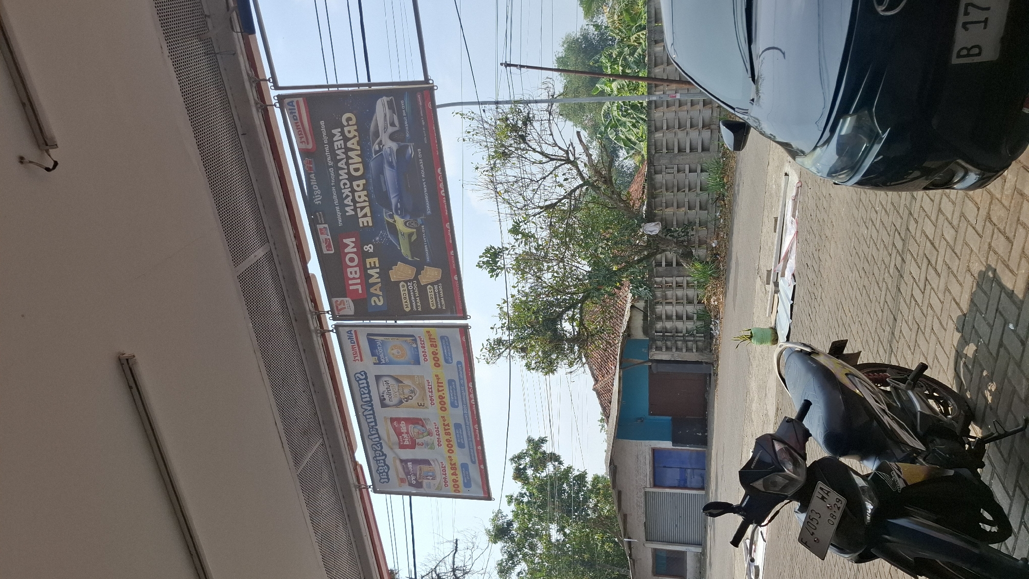 The image captures an outdoor scene in Cieundeur, Indonesia, on a bright, sunny day. Dominating the upper portion of the frame are two large advertising banners suspended from an overhang. The banner on the left displays images of milk powder products, each with a price listed as "000.825" and "000.625". The banner on the right is for a "GRAND PRIZE" promotion, featuring illustrations of cars and the text "MENANGKAN MOBIL & EMAS" (Win a Car & Gold).
In the foreground, a black motorcycle is parked on a paved walkway. The motorcycle's license plate is visible and reads "F 2053 WA 08.29". To the right of the motorcycle, the front end of a dark-colored car is visible.
The background reveals a residential or commercial area with buildings and greenery. A street runs behind the paved walkway, and a row of low-lying buildings with dark doors and windows lines the street. Beyond the buildings, a concrete wall with a repetitive pattern stretches across the scene, and trees with dense foliage are scattered throughout. The sky is clear and blue, indicating fair weather. The overall impression is a typical street view in a semi-urban Indonesian setting.