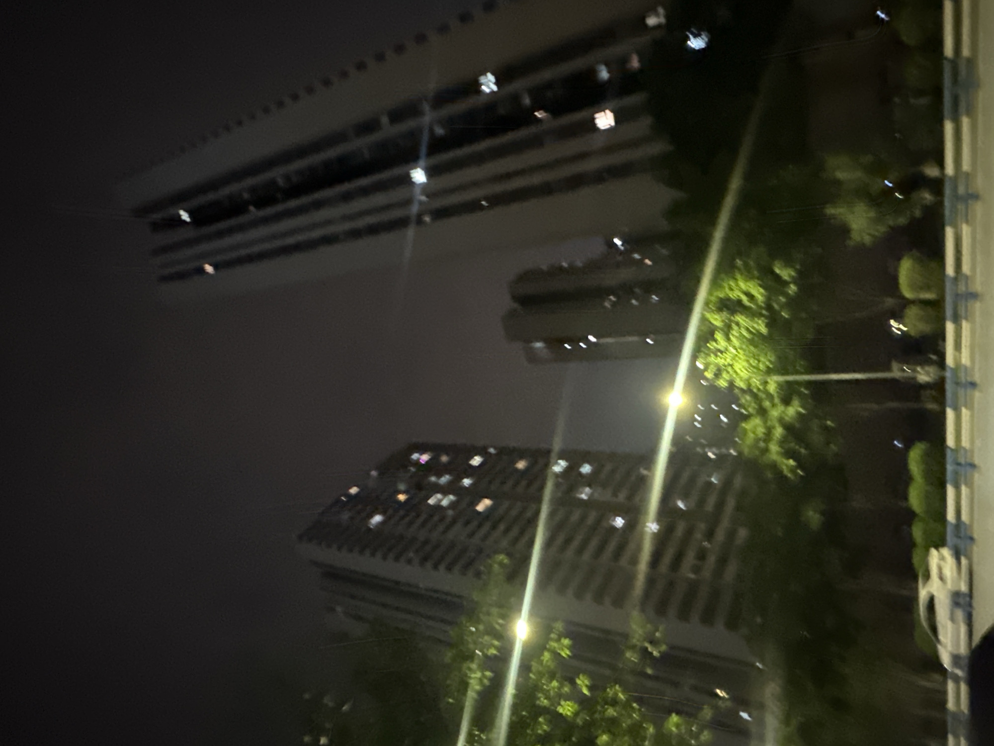 This media file depicts a nighttime scene in the Chongqing High-tech Zone, China. The primary subjects are two modern buildings, likely apartment complexes or offices, with multiple illuminated windows visible. The reflections of these buildings are visible on a dark, reflective surface, possibly water or a very smooth ground, suggesting the image might be taken at an unusual angle or that there is a reflection of the scene.
The setting appears to be an urban environment at night. Green trees and foliage are present in the foreground and between the buildings, indicating some form of landscaping or park area within the urban landscape. Several streetlights or building lights are visible, casting a glow and creating streaks of light in the image due to the long exposure or motion blur.
The activity taking place is not explicitly discernible, but the illuminated windows suggest that people are inside the buildings, possibly working or living. The overall mood is serene and perhaps a bit mysterious due to the nighttime setting and reflections. There is no visible text in the image. The weather appears to be clear, as there are no indications of rain or fog. The time of day is clearly night.