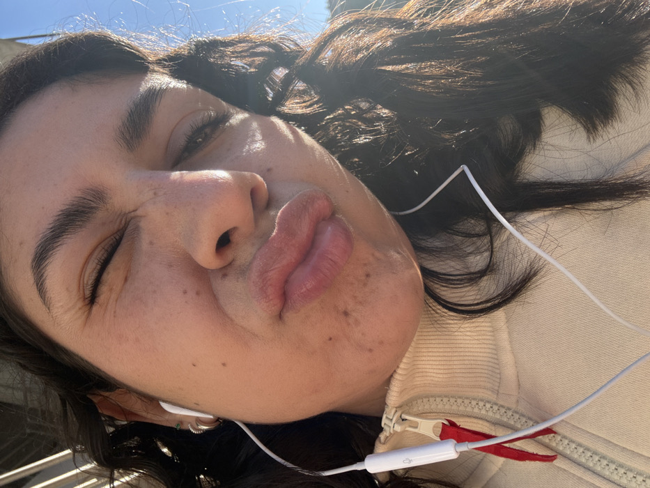 A close-up, angled selfie shows a young woman's face, partially obscured by strong, bright sunlight. Her eyes are closed or squinted, and her lips are puckered in a "duck face" expression, conveying a playful mood. Her dark hair frames her face, and her skin has visible freckles. She is wearing a cream or light-colored hooded jacket with a zipper, and white wired earphones are draped over her. A small control unit on the earphone cable, featuring an indistinct icon (likely for microphone/volume), is visible against her jacket. The background shows a clear blue sky, indicating bright, sunny weather, likely during midday or early afternoon given the intense light. The setting appears to be outdoors in Hialeah, United States of America.