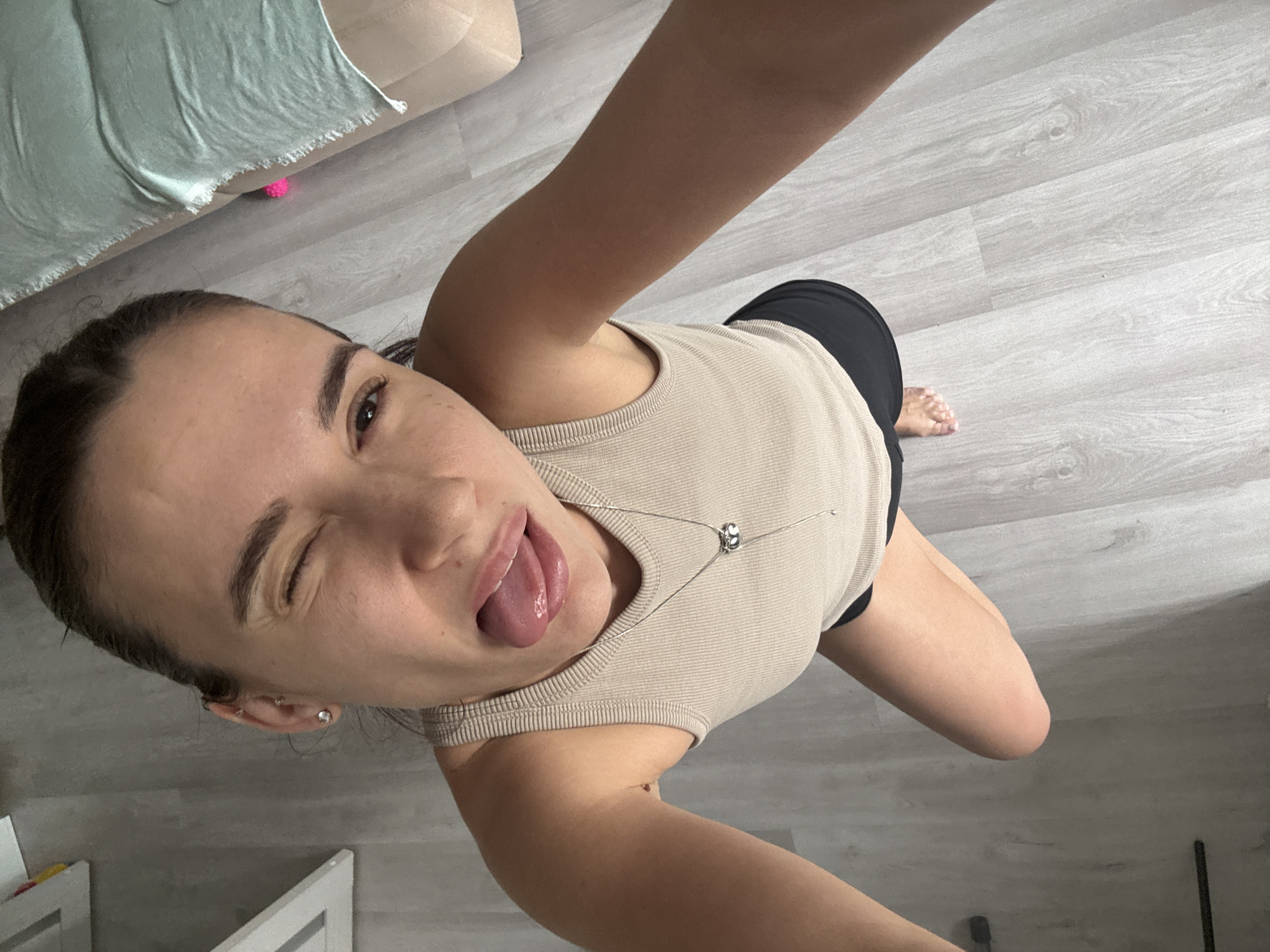 A woman takes a selfie with an open mouth and her tongue sticking out, while winking. She wears a light-colored ribbed tank top with a silver necklace, and black shorts. She stands on a light gray wood-look floor. A pink ball and part of a green blanket rest on the floor in the background, near a light beige sofa. The location appears to be indoors in a home. There is no information in the image to confirm the location is in the city of Chaikovsky, Russia.